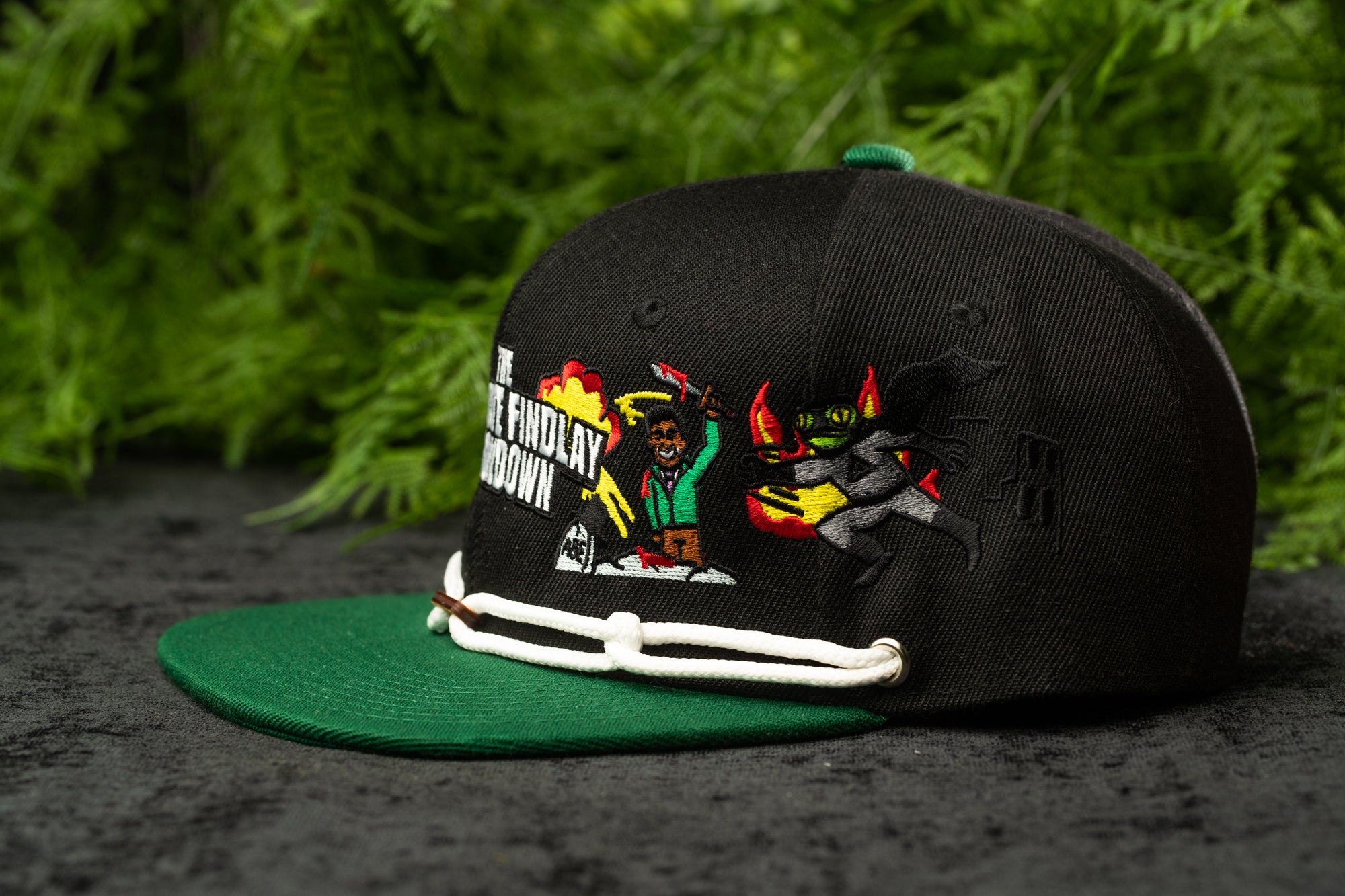 A black baseball cap with a green brim and a colorful graphic design on the front, featuring a cartoon character and text.