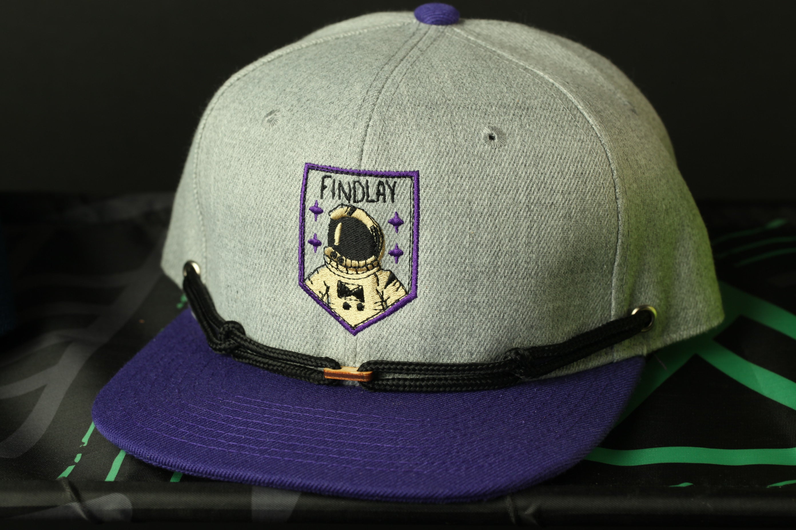 A gray baseball cap with a purple brim and a purple and white patch on the front, featuring an astronaut helmet and the word "FINDLAY" written on it.