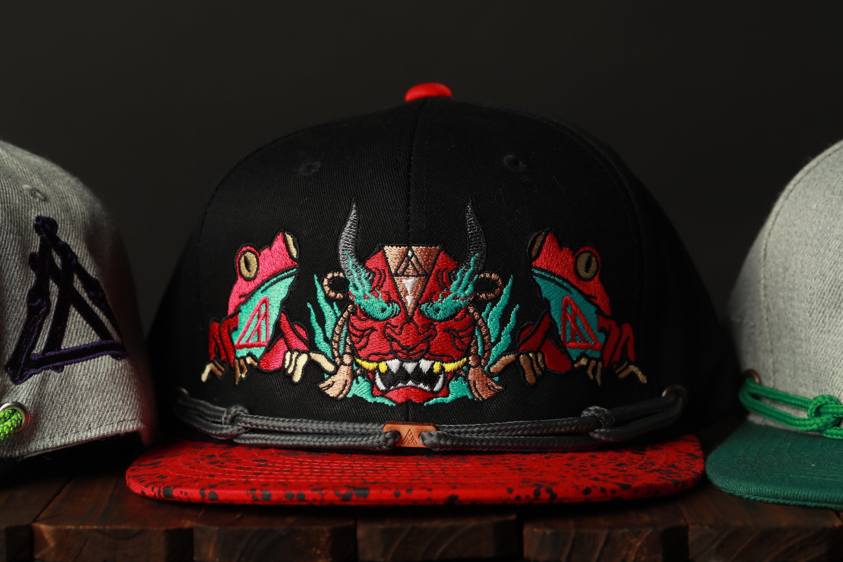 A close-up view of a black baseball cap with colorful frog designs on it, placed on a wooden surface.