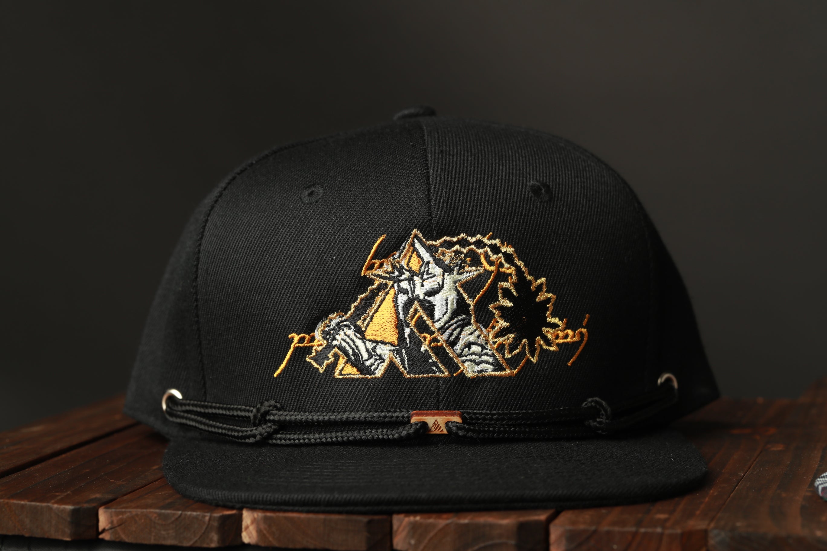 A black baseball cap with an embroidered design on the front, sitting on a wooden surface.