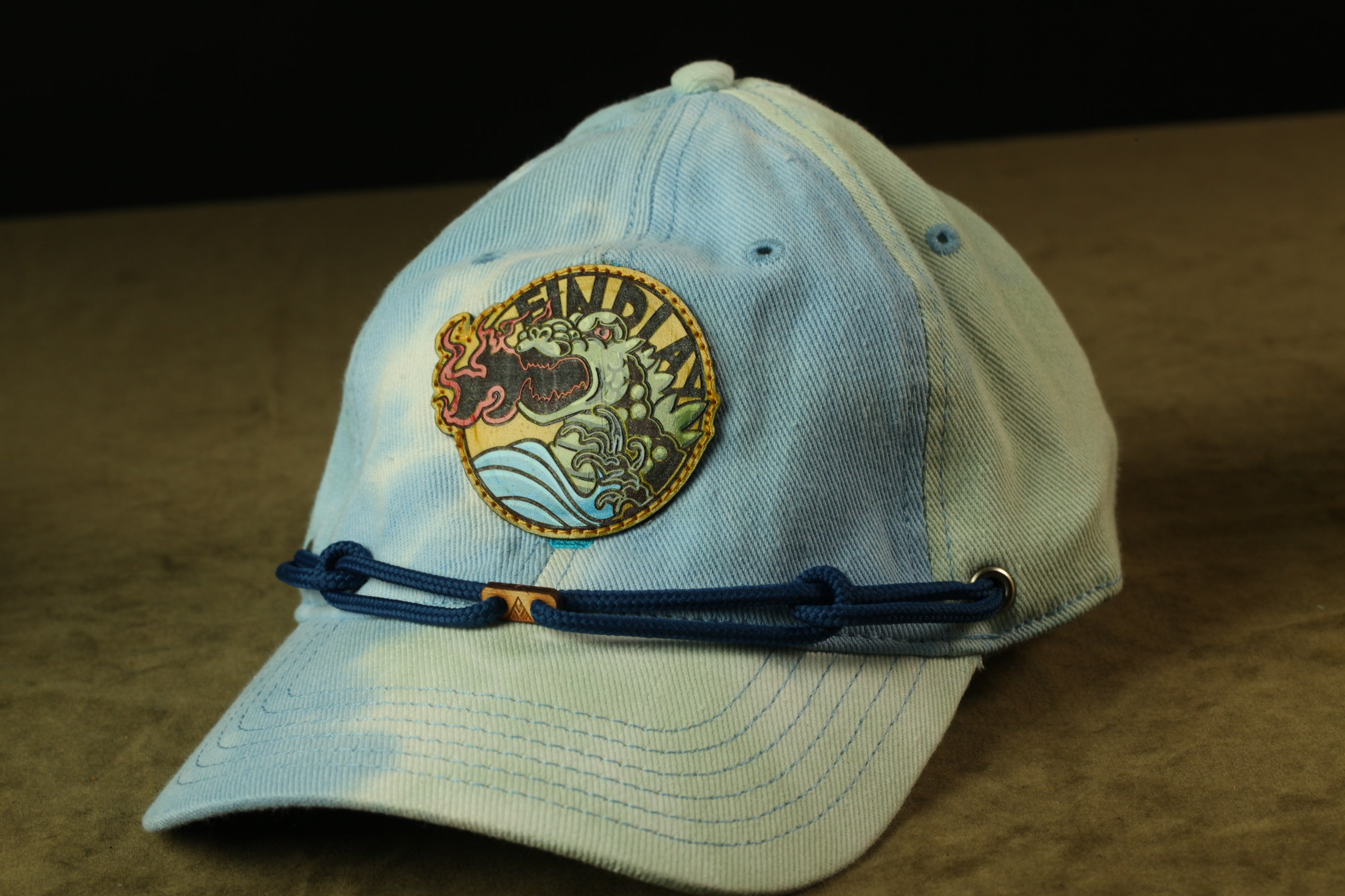 A light blue baseball cap with a circular patch on the front, featuring a dragon and waves, is shown on a brown surface.