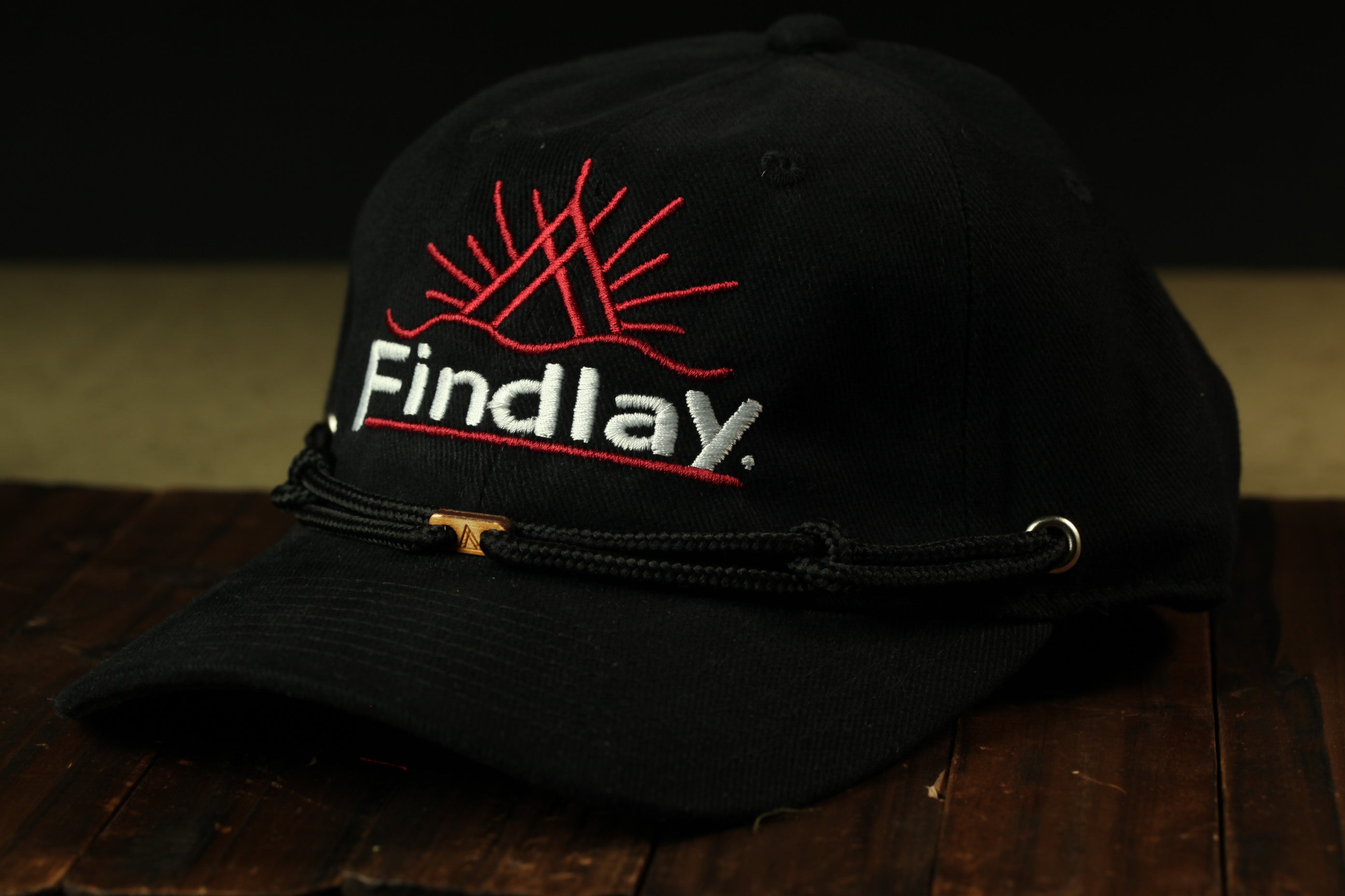 A black baseball cap with a red sun design and the word "Findlay" printed on it, sitting on a wooden surface.