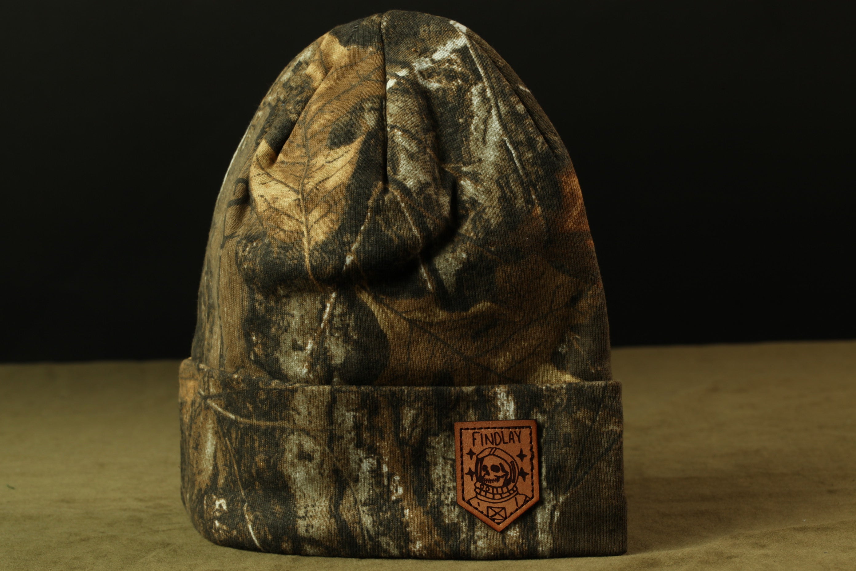 A camouflage-patterned beanie with a patch on the front displaying the text "INDOLAY".