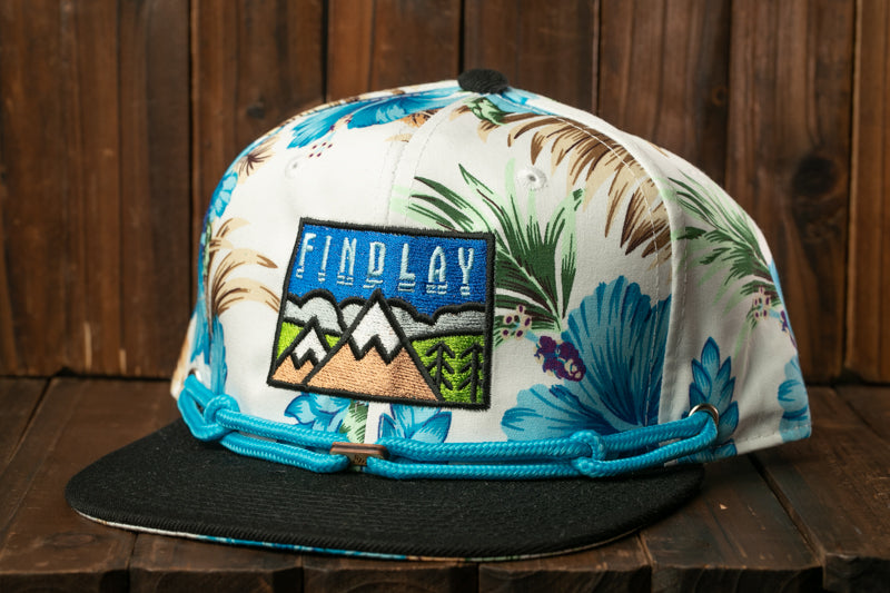 A white baseball cap with a floral print and a patch that says "FIND LAY" is displayed on a wooden surface.
