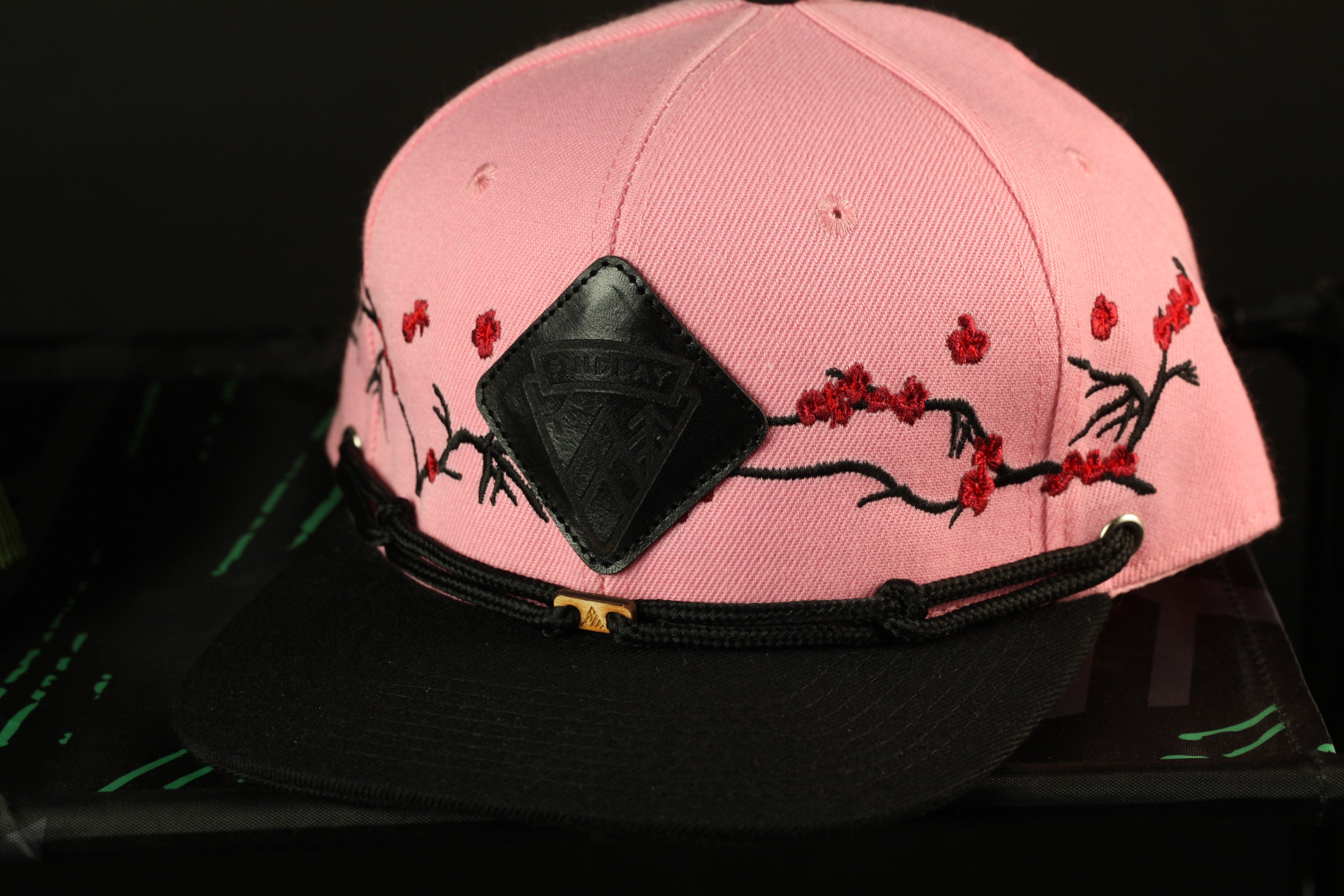 A pink baseball cap with a black band and a black and red floral embroidered design on the front.