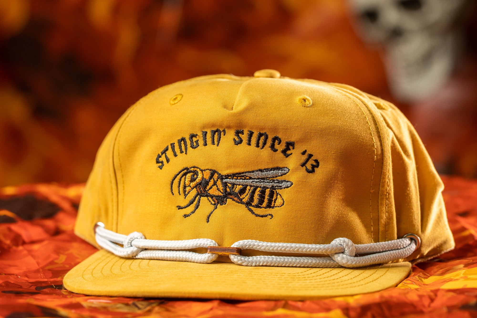 A yellow baseball cap with a black and white bee embroidered on it, sitting on a red and orange patterned fabric.