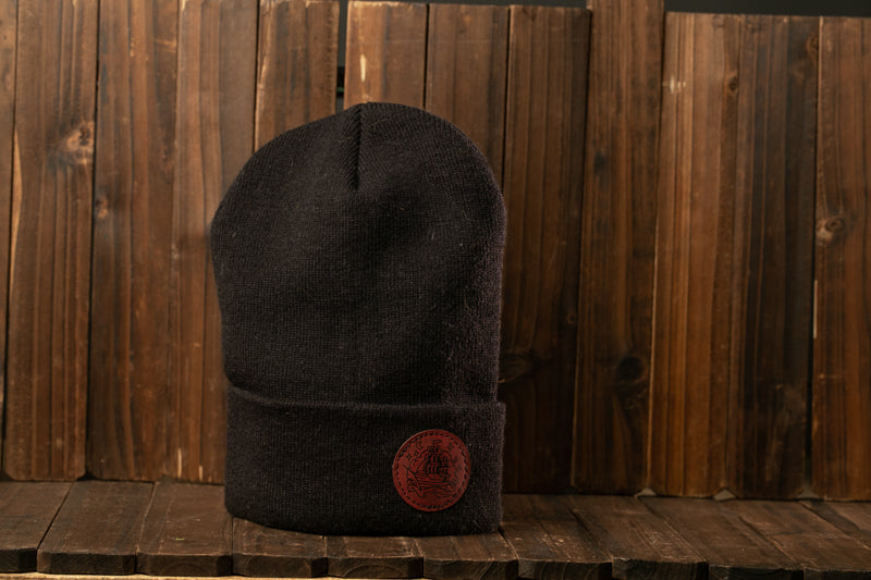 A black knit cap with a red circular logo is displayed against a wooden background.