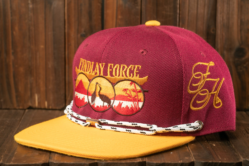 A maroon baseball cap with a yellow brim and a white rope strap, featuring embroidered designs and text.