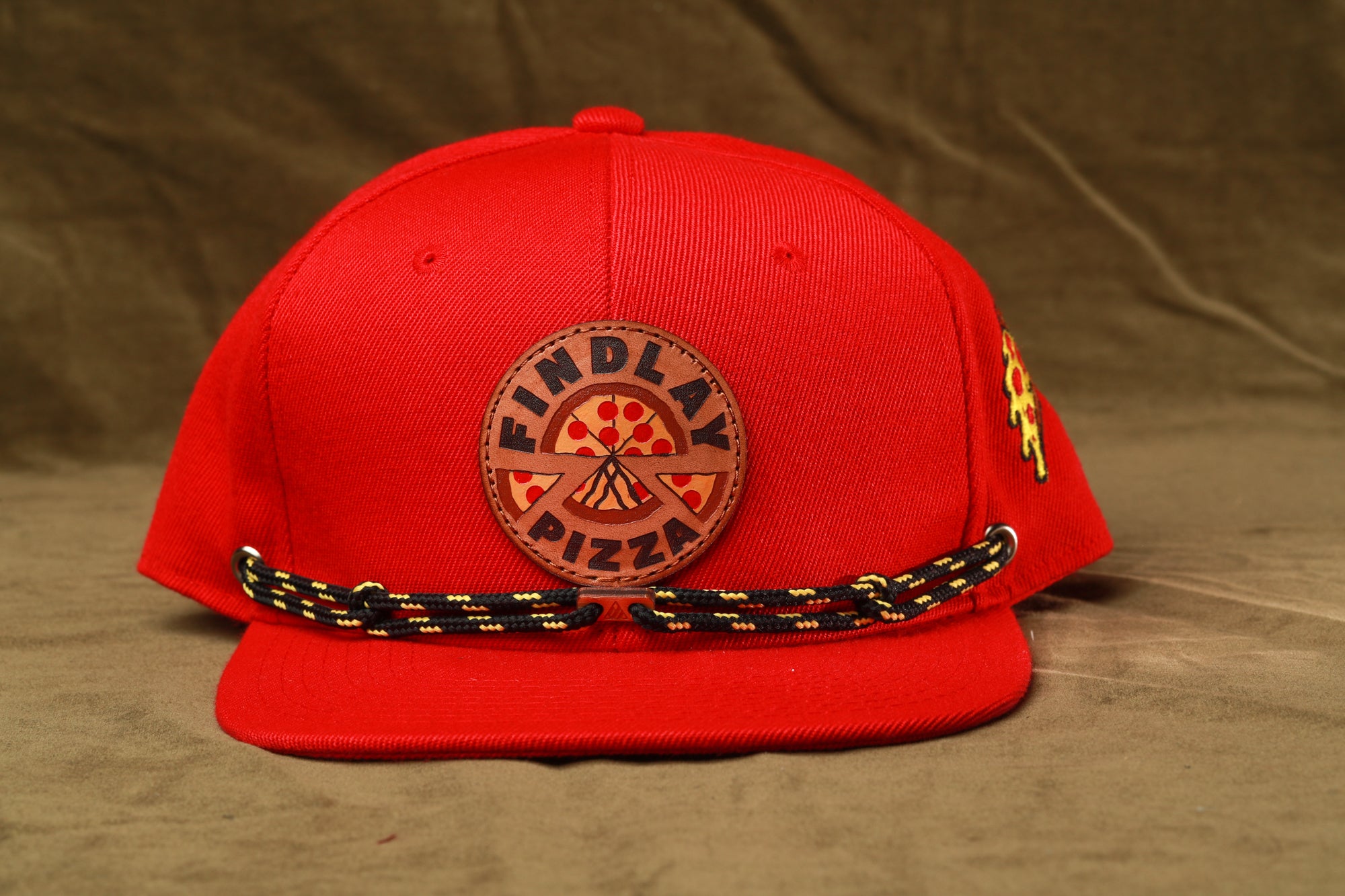 A red baseball cap with a black and yellow rope strap and a circular logo on the front, which appears to be a logo for a pizza restaurant.