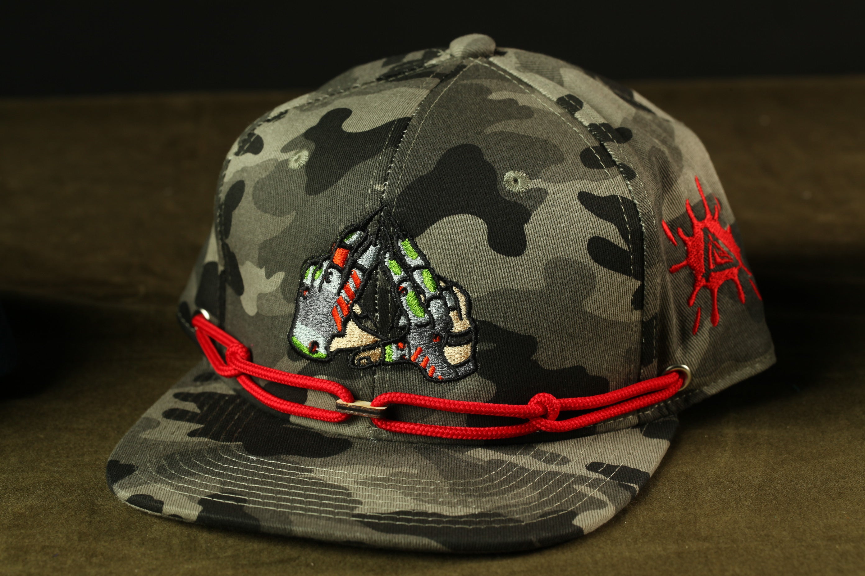A camouflage baseball cap with a red chain strap and a graphic design on the front.