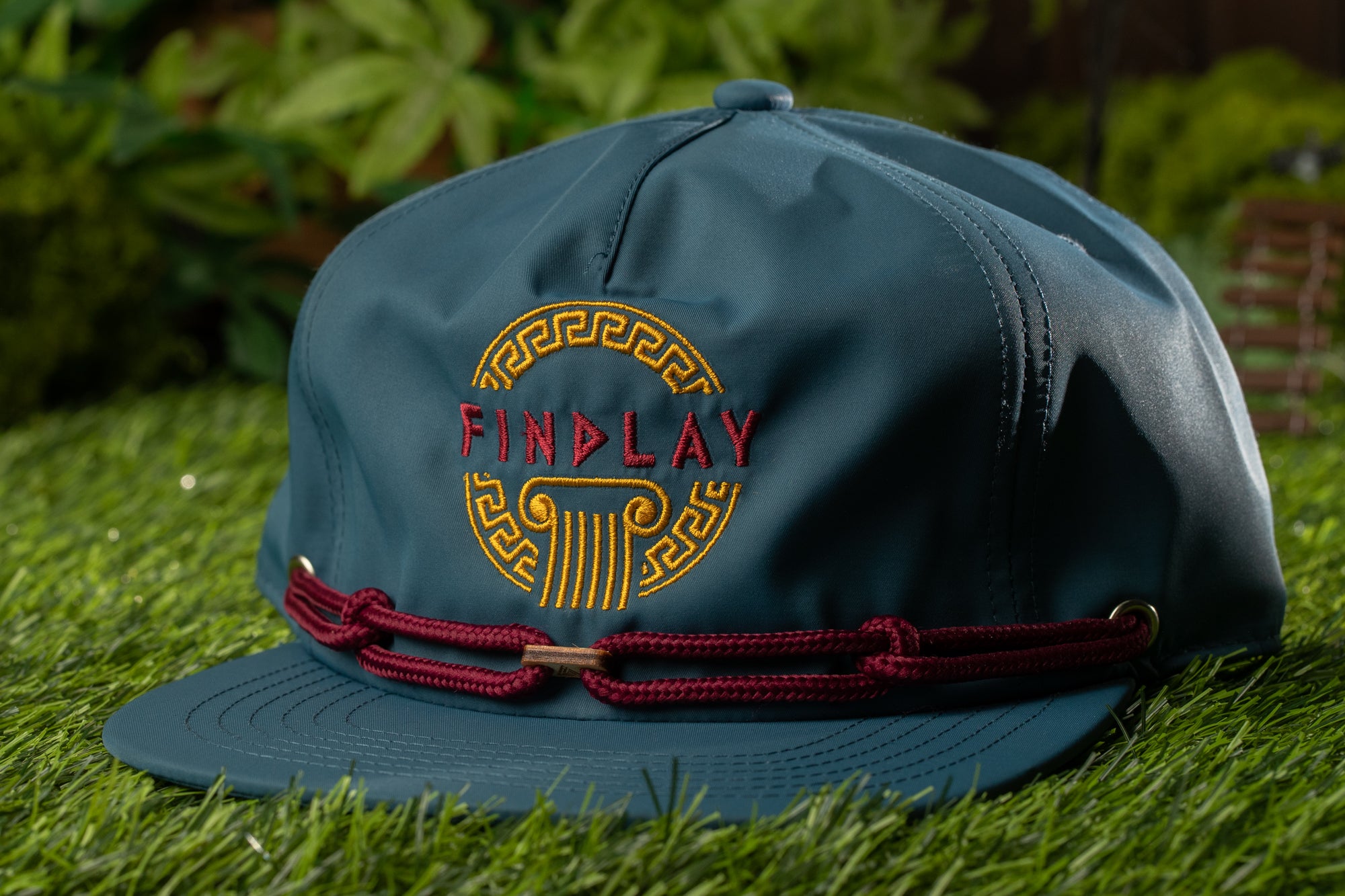 A dark green baseball cap with a maroon band and the word "FINDPLAY" embroidered on it, placed on a grassy surface.