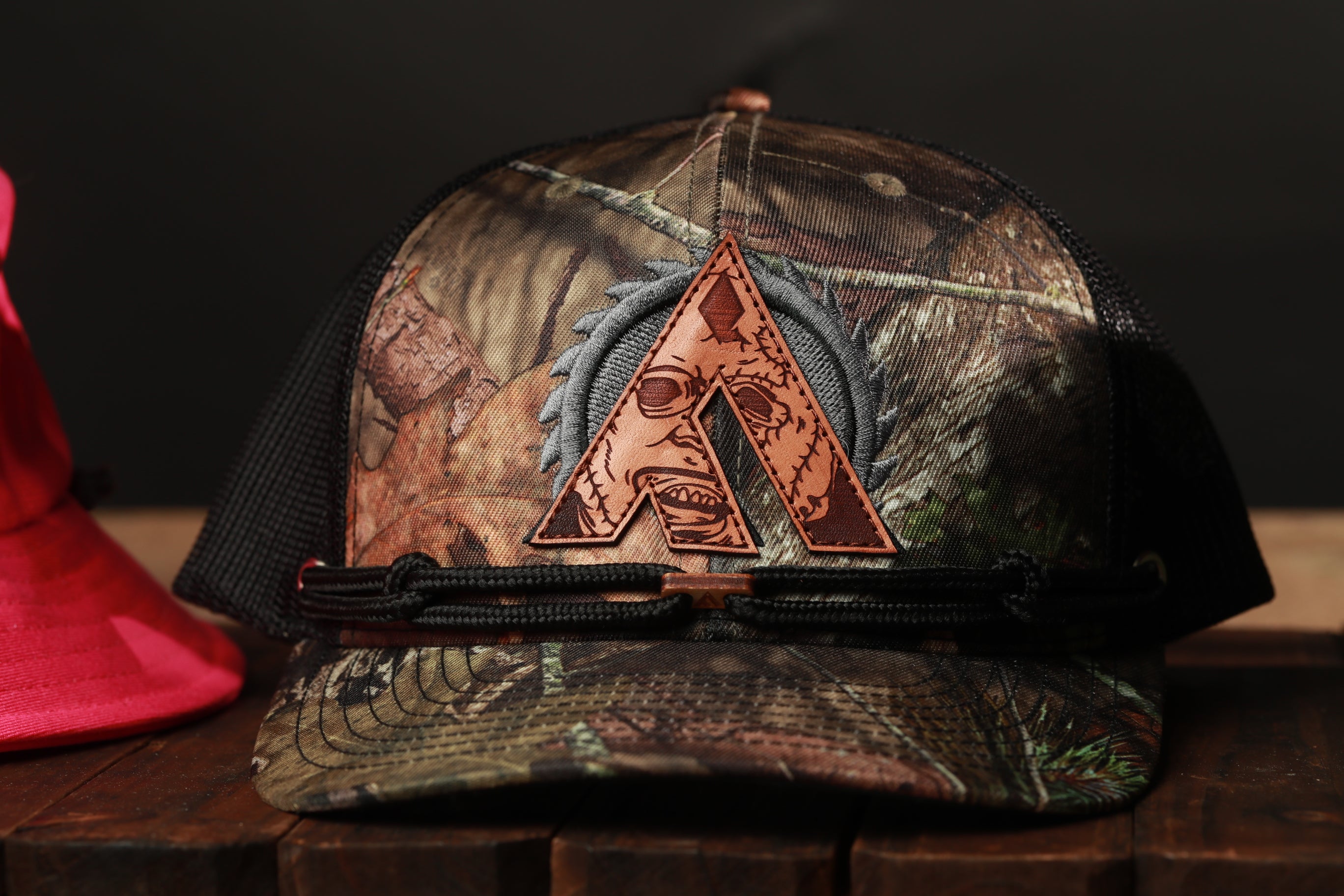 A camouflage-patterned baseball cap with a leather patch featuring a stylized letter 'A' on it, placed on a wooden surface.