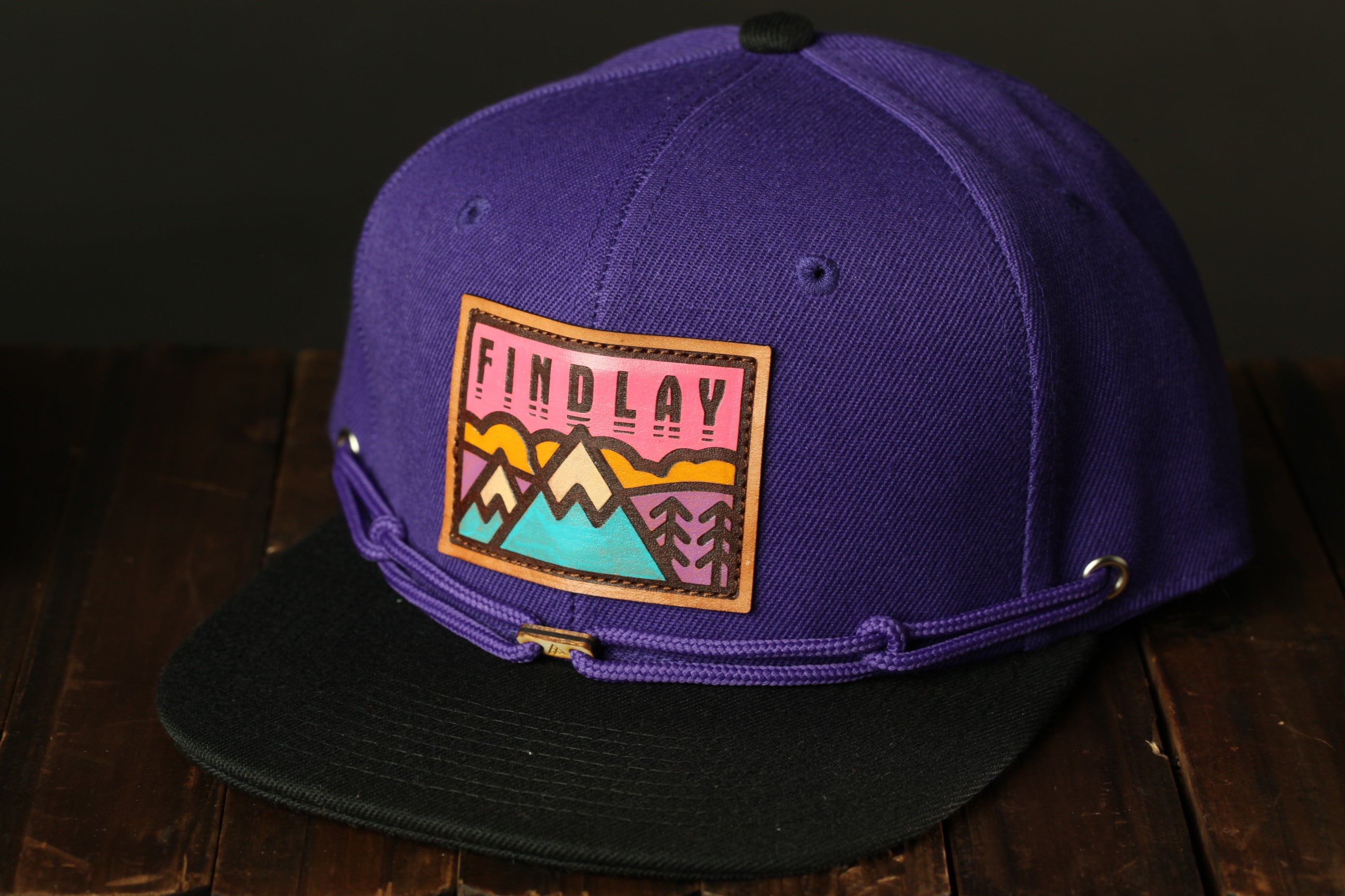 A purple baseball cap with a colorful patch on the front, featuring a mountain landscape design.