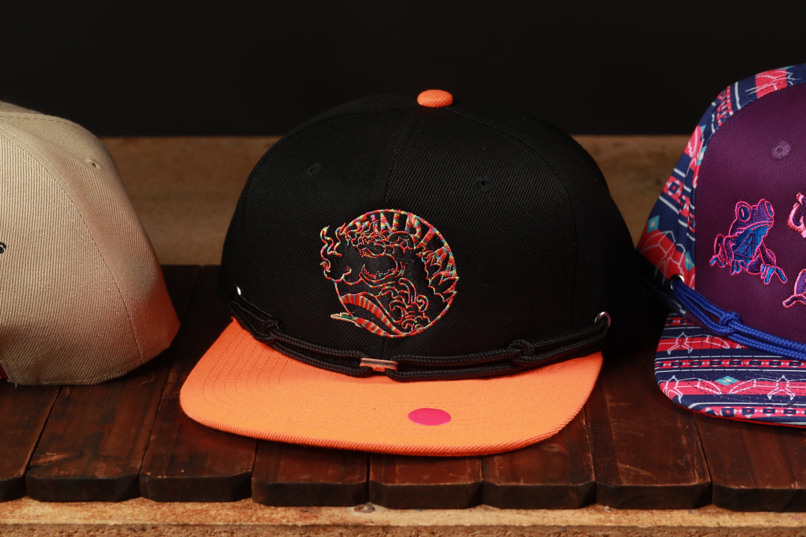 Three baseball caps are displayed on a wooden surface, with one black cap featuring a colorful graphic and two other caps in different colors and designs.
