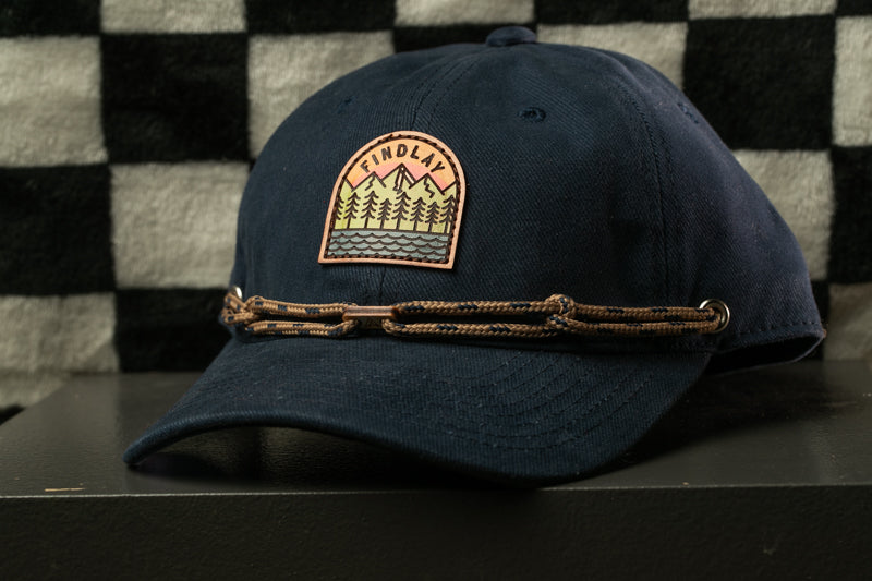 A navy blue baseball cap with a patch on the front, featuring a mountain landscape design, is displayed on a black surface against a checkered background.