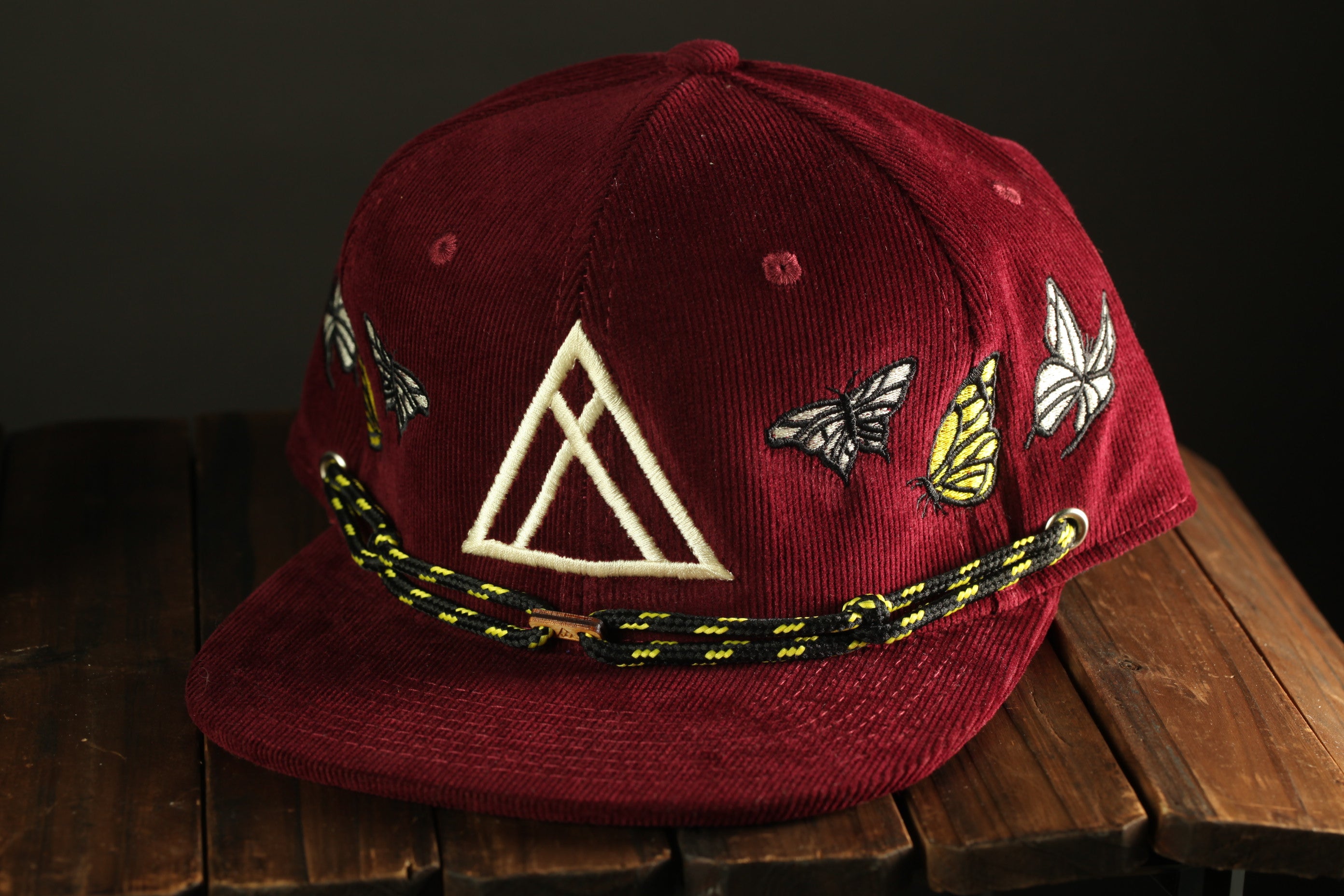 A burgundy baseball cap with a white triangle and colorful butterfly designs on the front, sitting on a wooden surface.