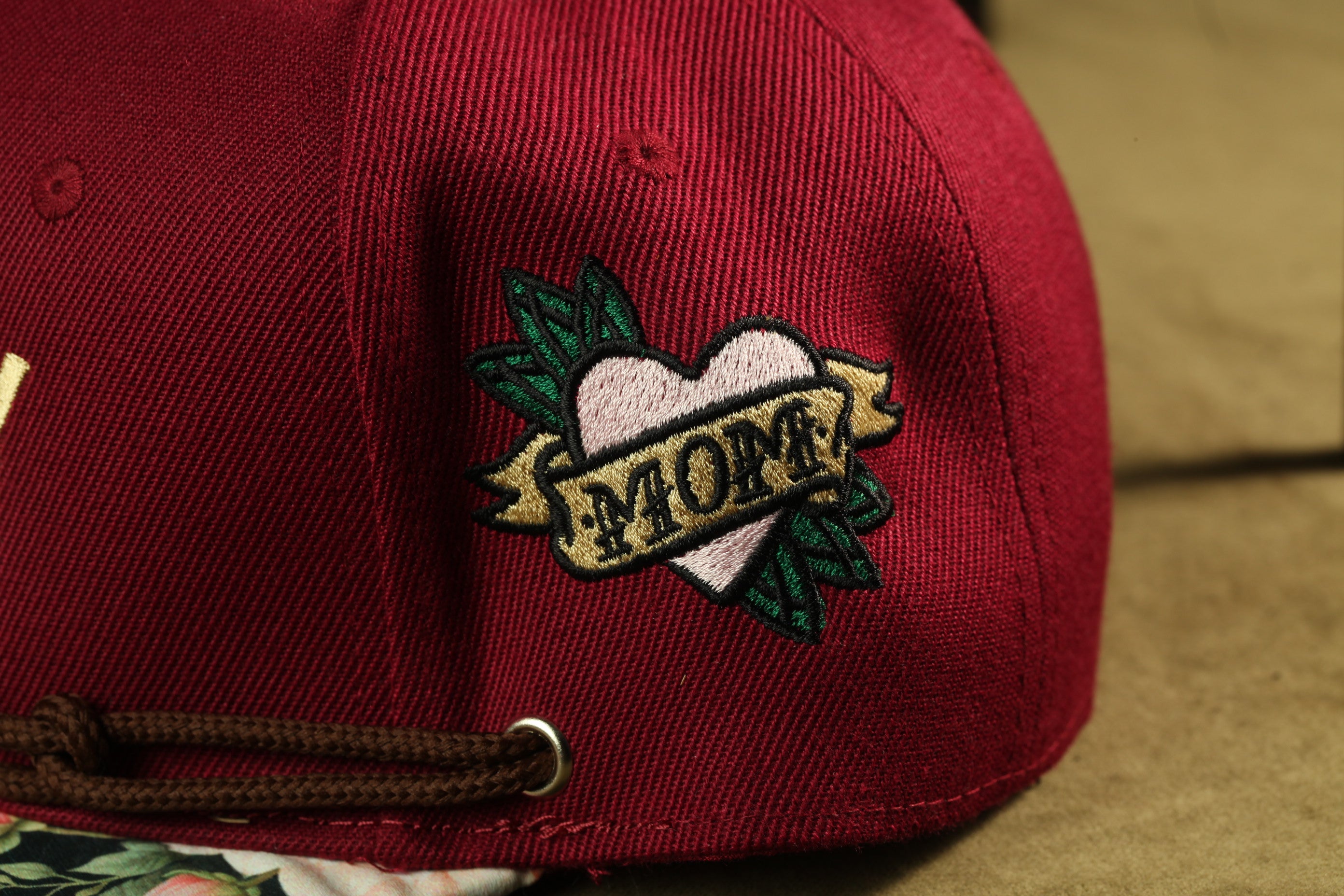 A close-up of a red baseball cap with a heart-shaped embroidered patch that says "MOM" on it.