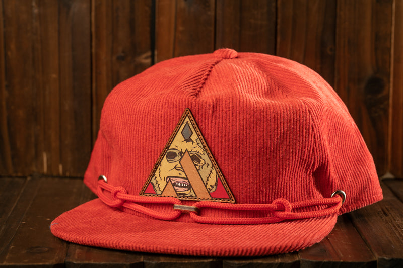 A red baseball cap with a triangular patch featuring a cartoon character is displayed on a wooden surface.