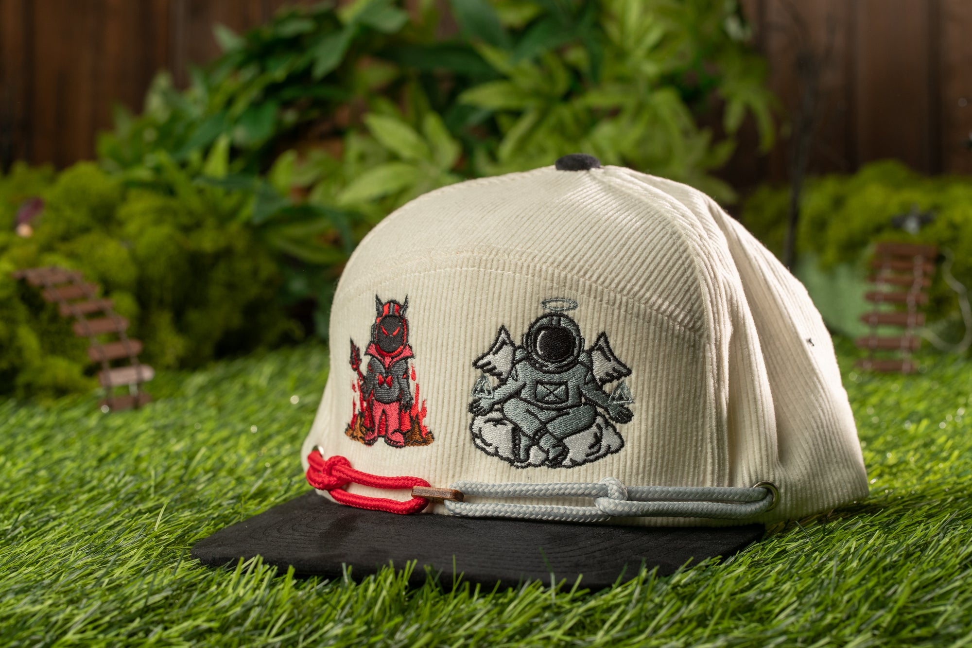 A beige baseball cap with two cartoon characters on it, one of which is an angel, is placed on a grassy surface.