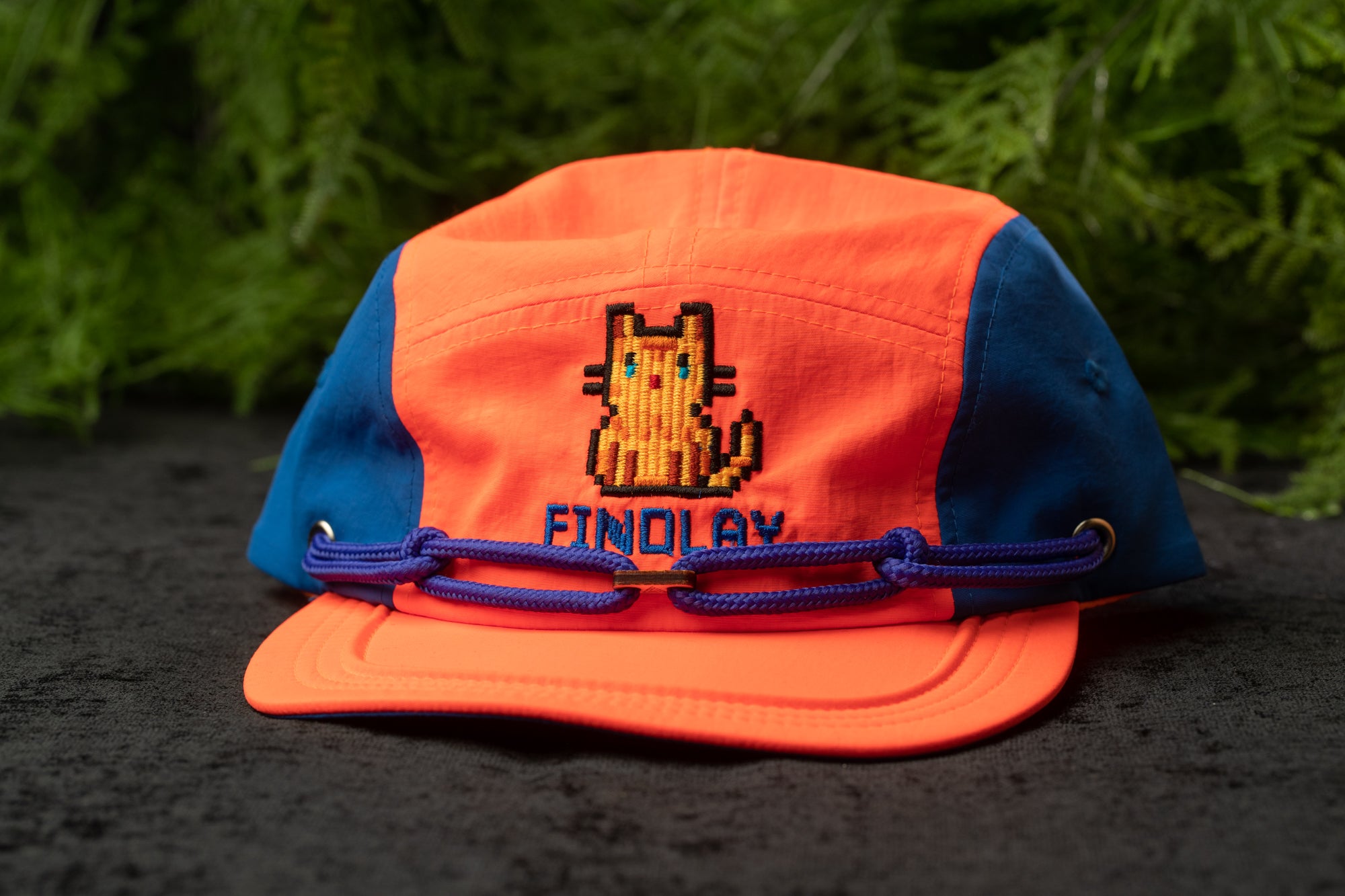 An orange and blue baseball cap with a cat embroidered on it, sitting on a black surface with greenery in the background.