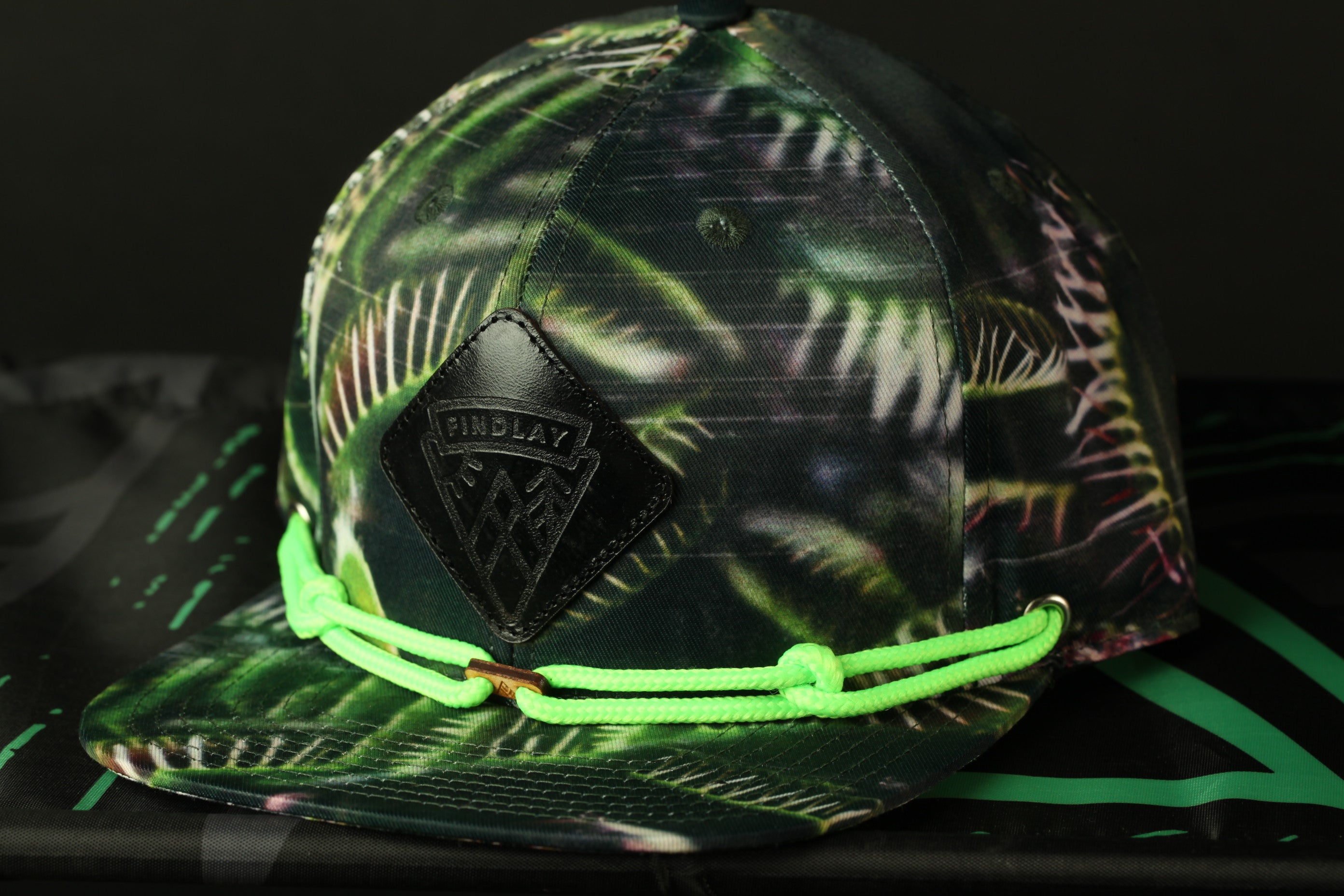 A camouflage-patterned baseball cap with a black leather patch and neon green strings.