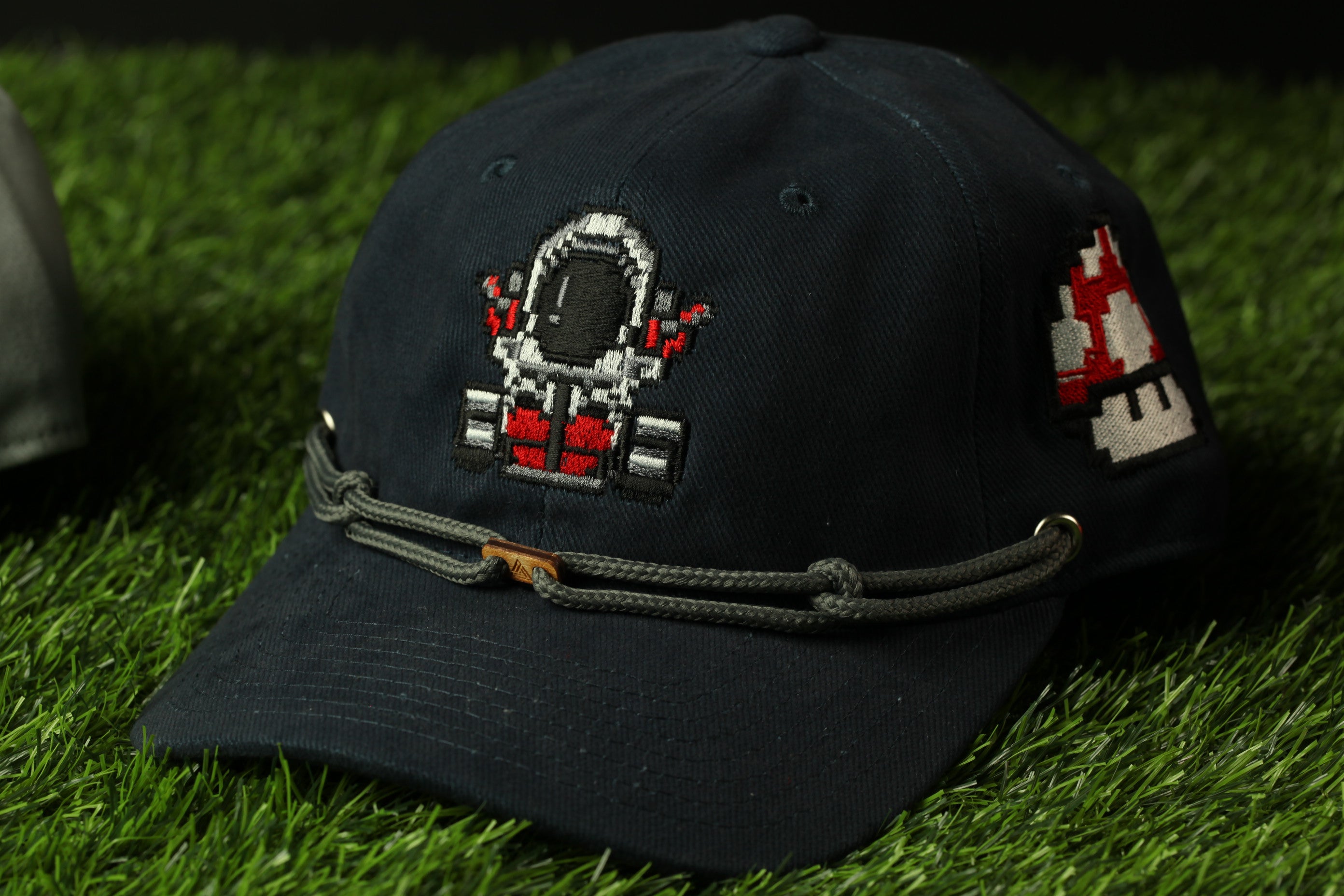 A black baseball cap with a pixelated astronaut design on the front, sitting on a grassy surface.