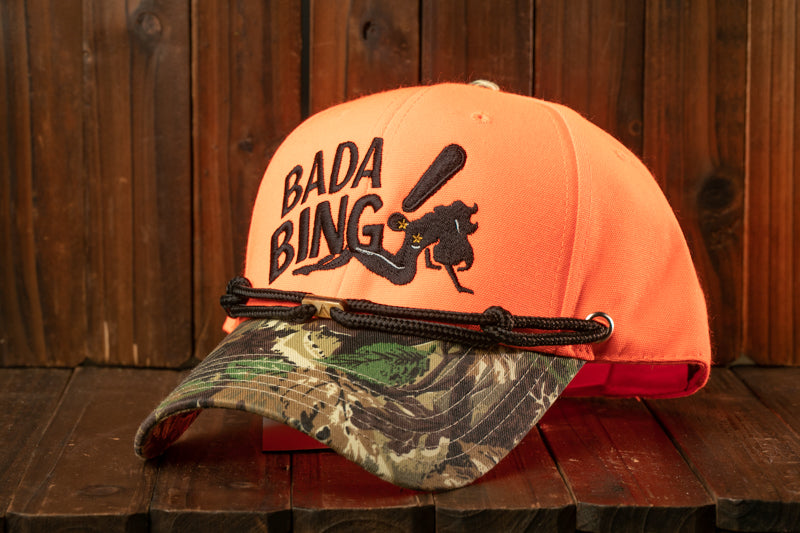 An orange baseball cap with the text "Bada Bing" printed on it, placed on a wooden surface.