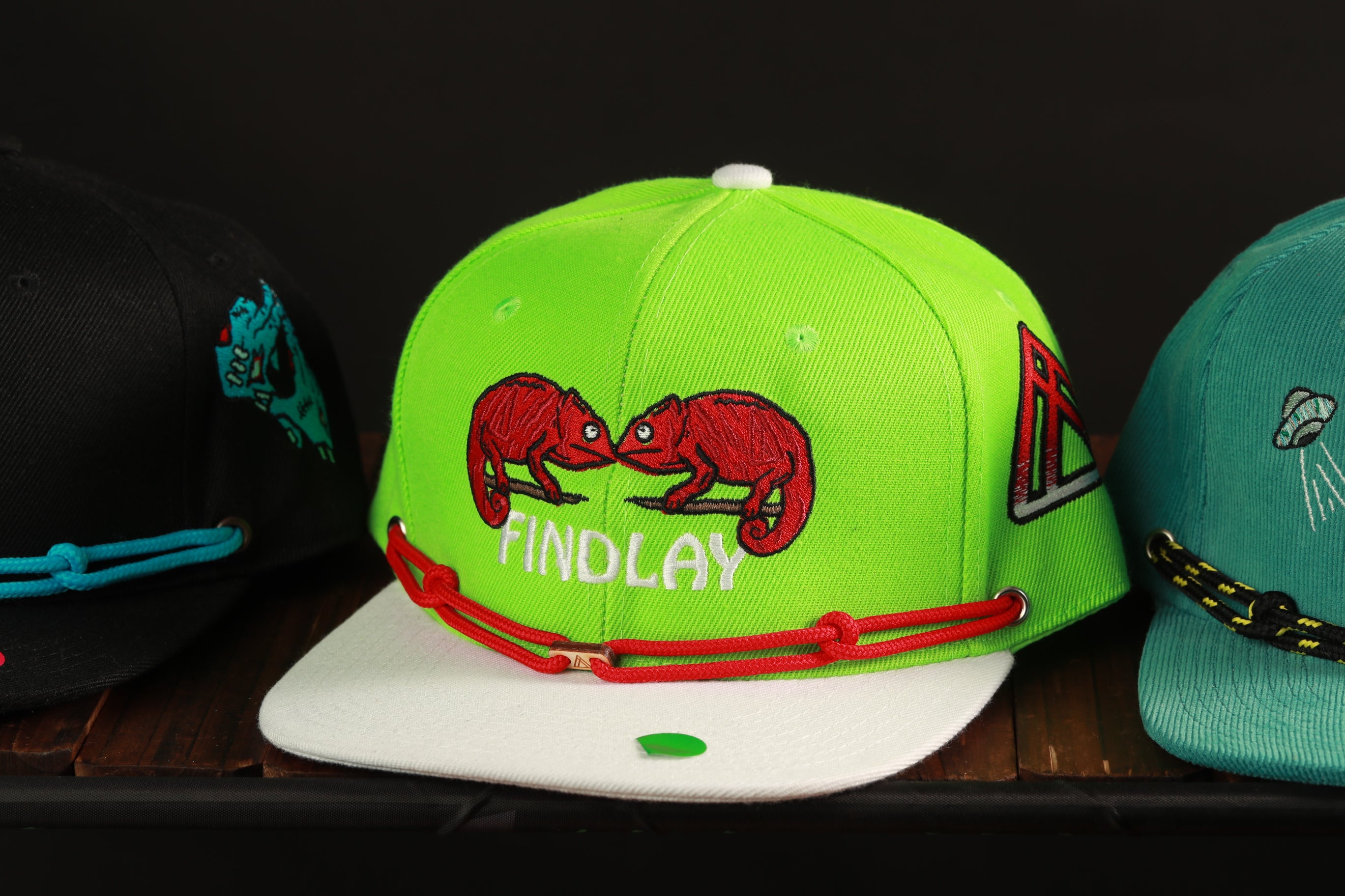 A collection of colorful baseball caps with various designs and colors, including a lime green cap with a red chameleon and the word "FINDLAY" printed on it.