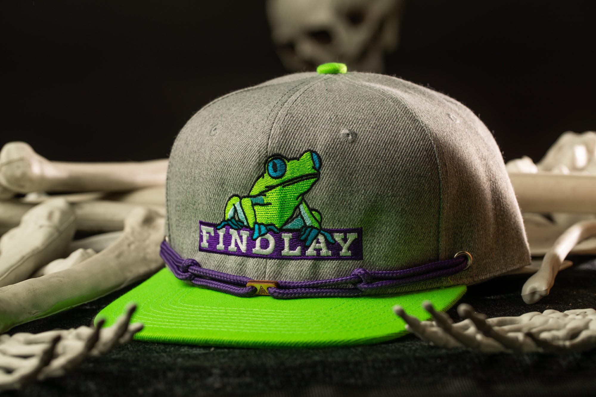 A gray baseball cap with a green frog design and purple text, placed on a surface with white bones.