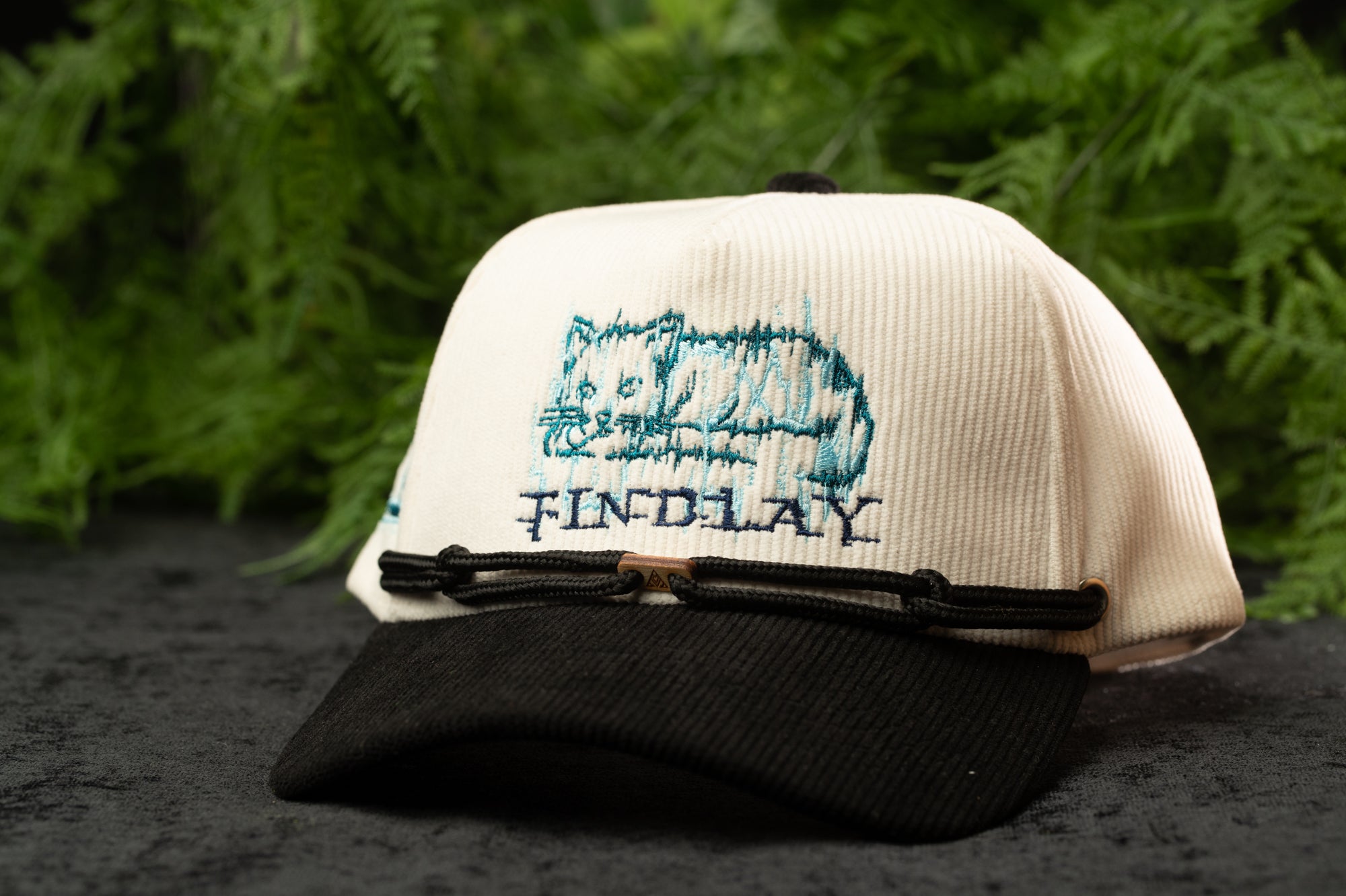 A white baseball cap with a cat embroidered on it, placed on a black surface with greenery in the background.
