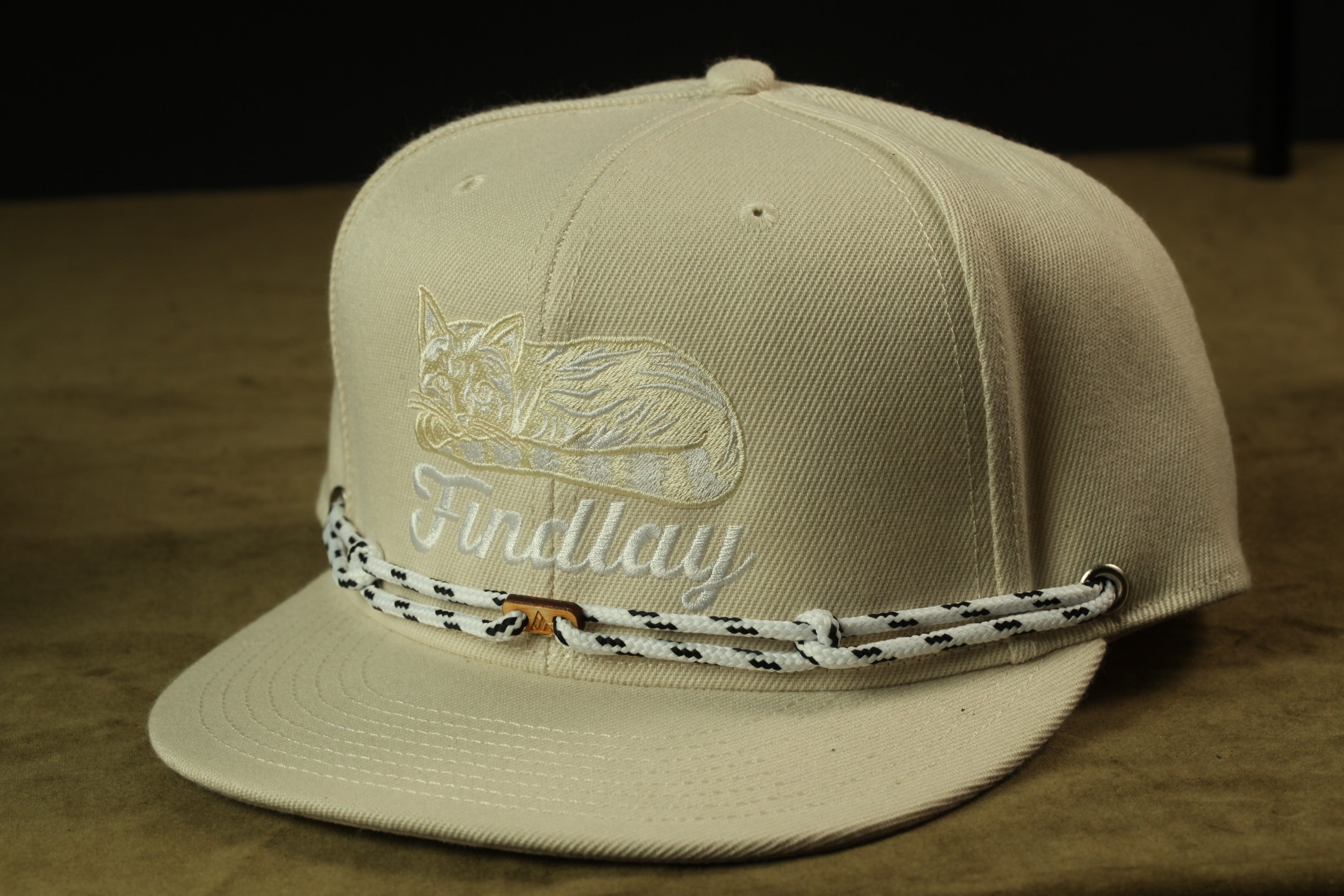 A beige baseball cap with a cat embroidered on it, and a black and white rope strap around the brim.