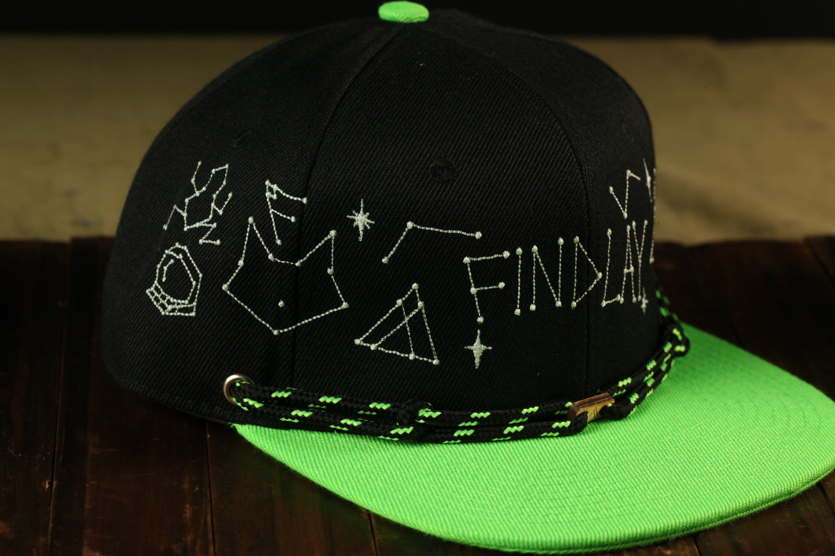 A black baseball cap with a green brim, featuring white embroidered designs and text on the front.