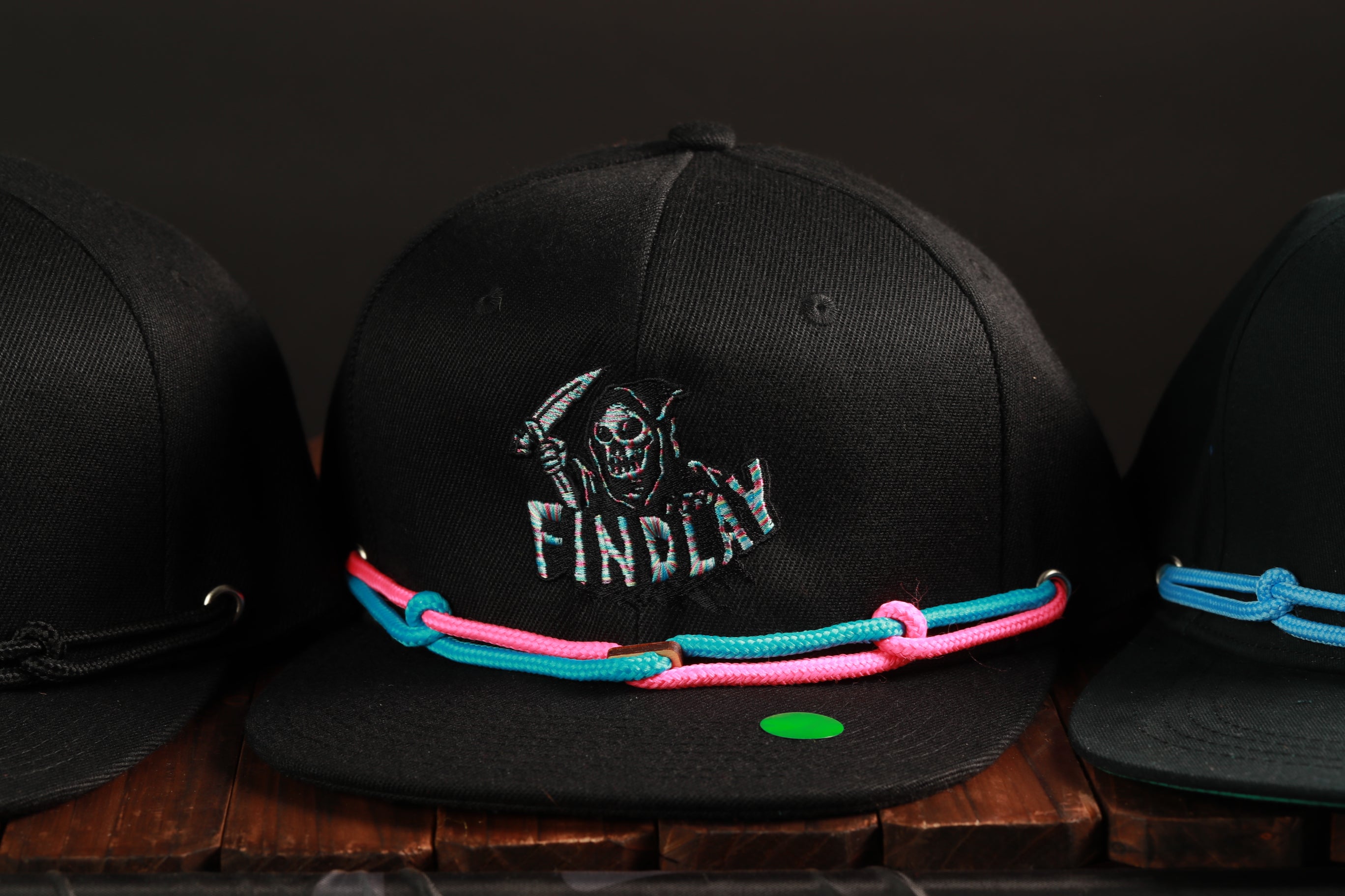 A black baseball cap with a skull and crossbones design and the word "FINDLAY" printed on it, along with a colorful braided strap.