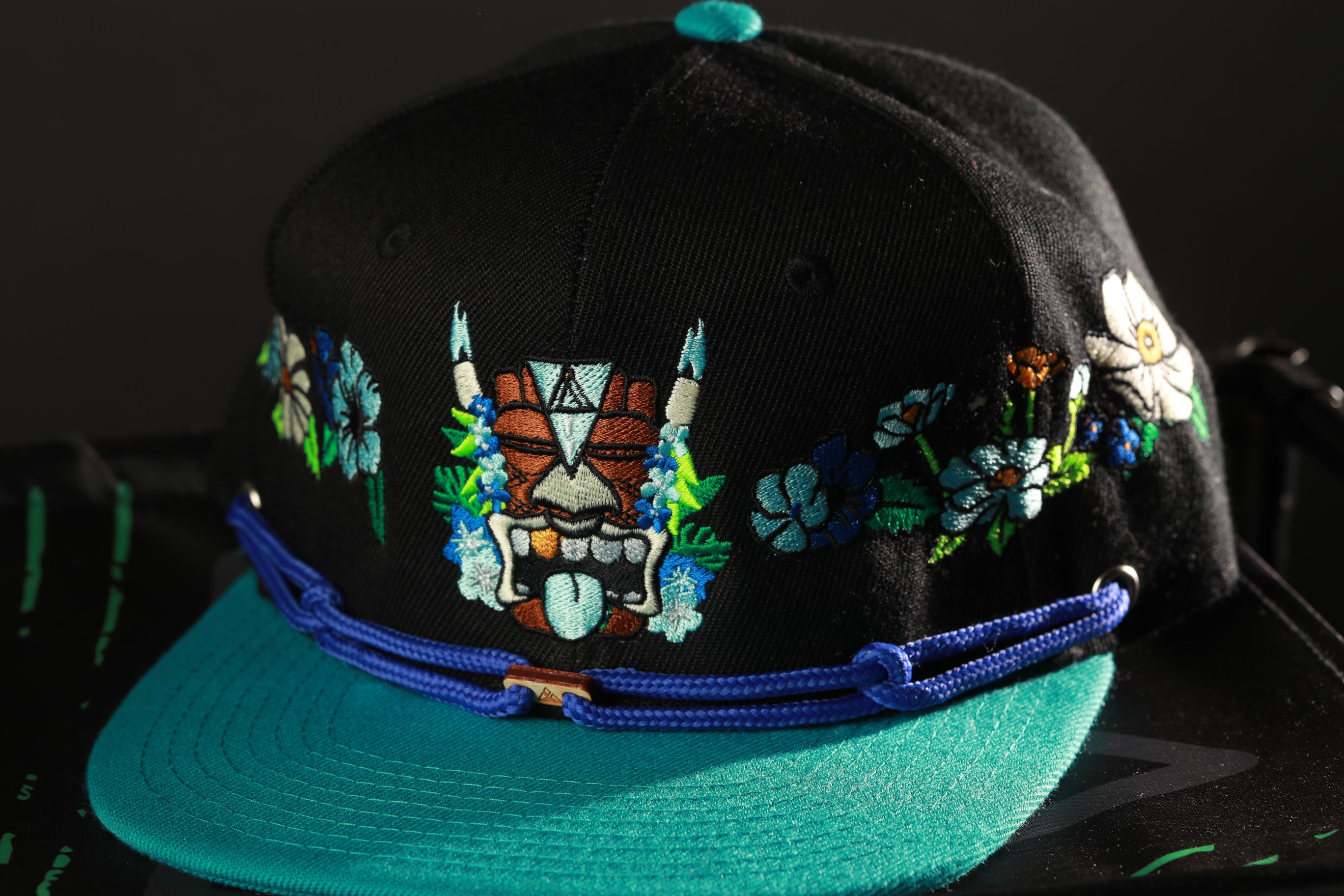 A black baseball cap with a teal brim and colorful floral embroidery on the front.