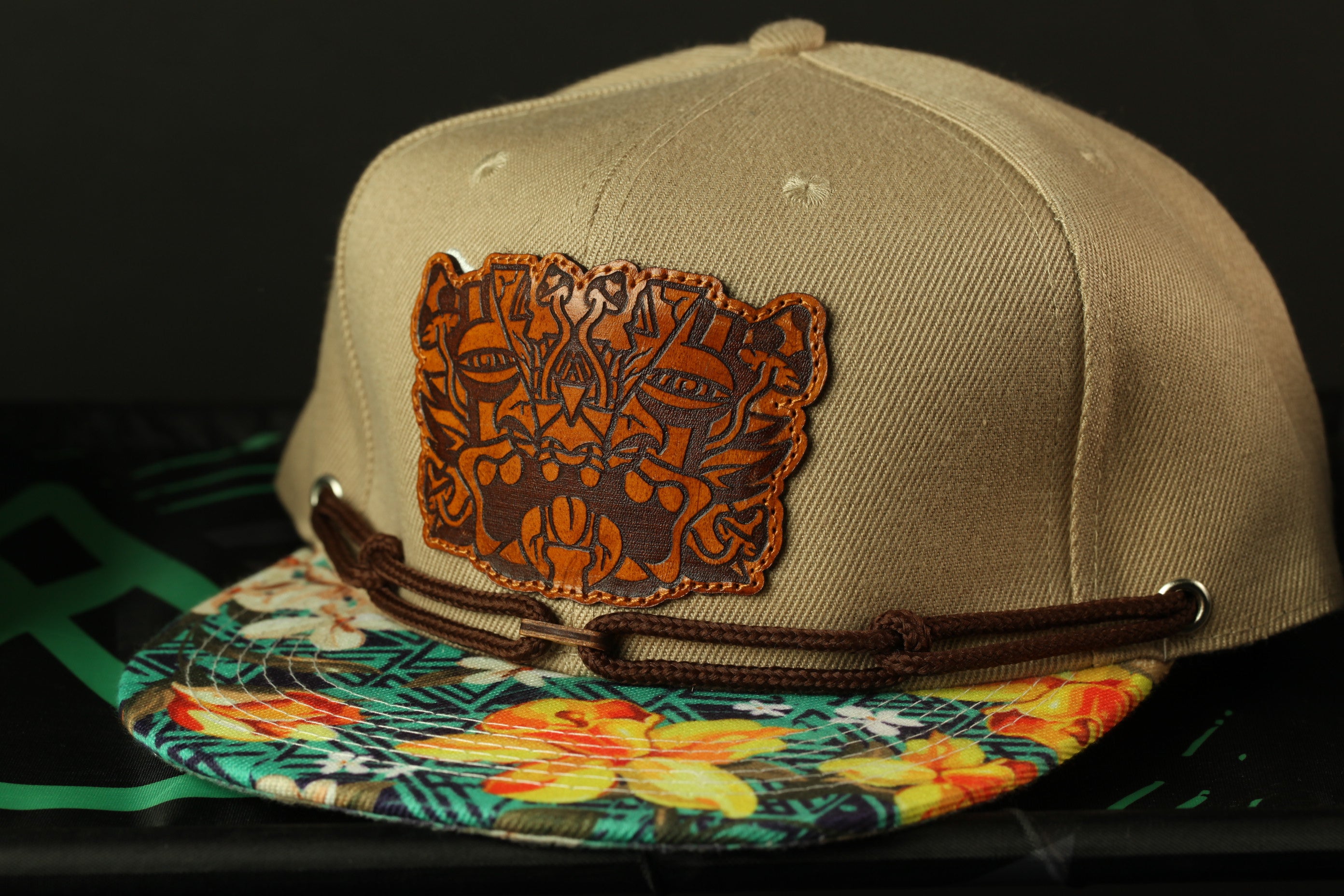 A beige baseball cap with a brown leather patch featuring a floral design, placed on a colorful printed fabric.