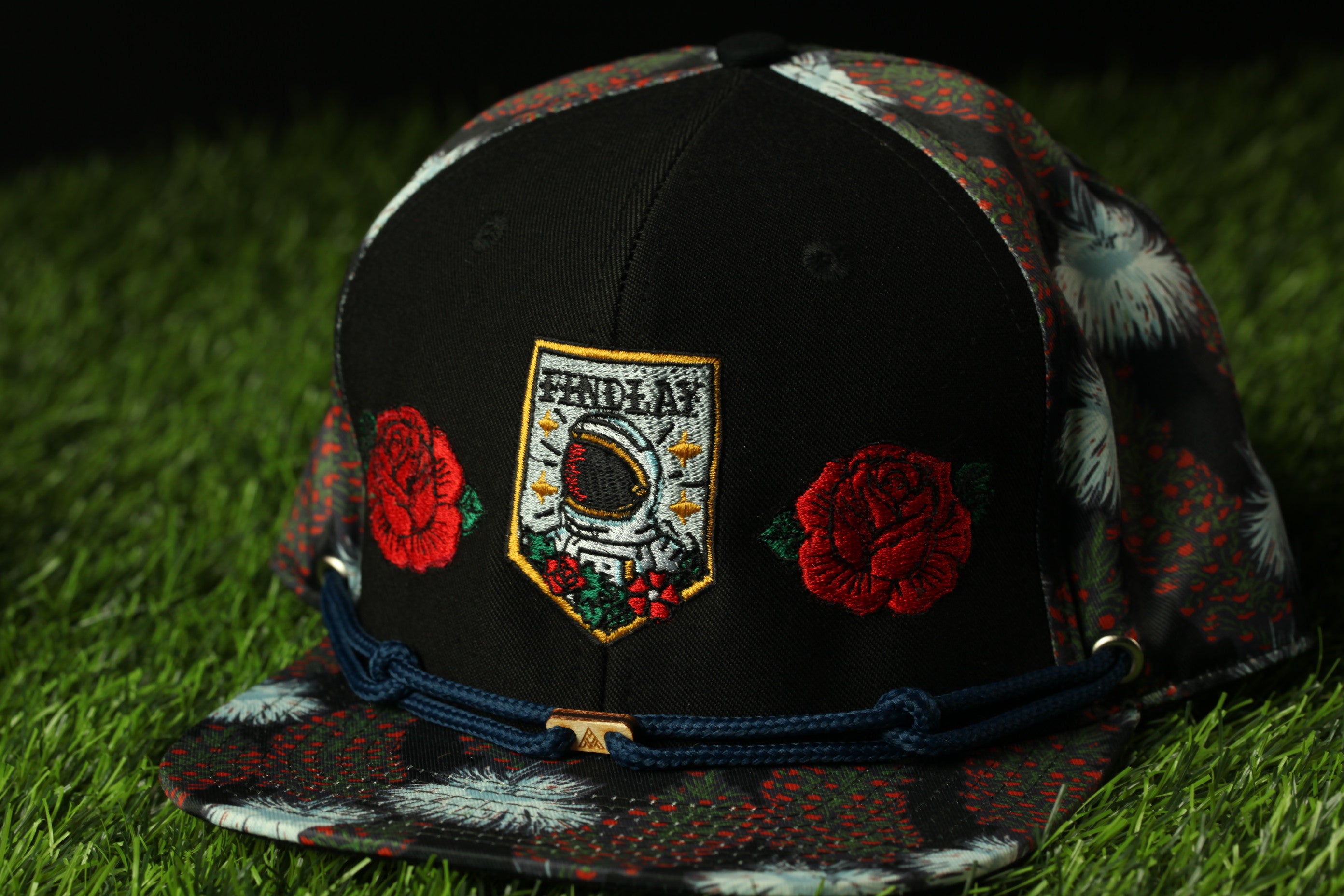 A black baseball cap with a floral pattern and a patch featuring a space shuttle design, resting on a grassy surface.