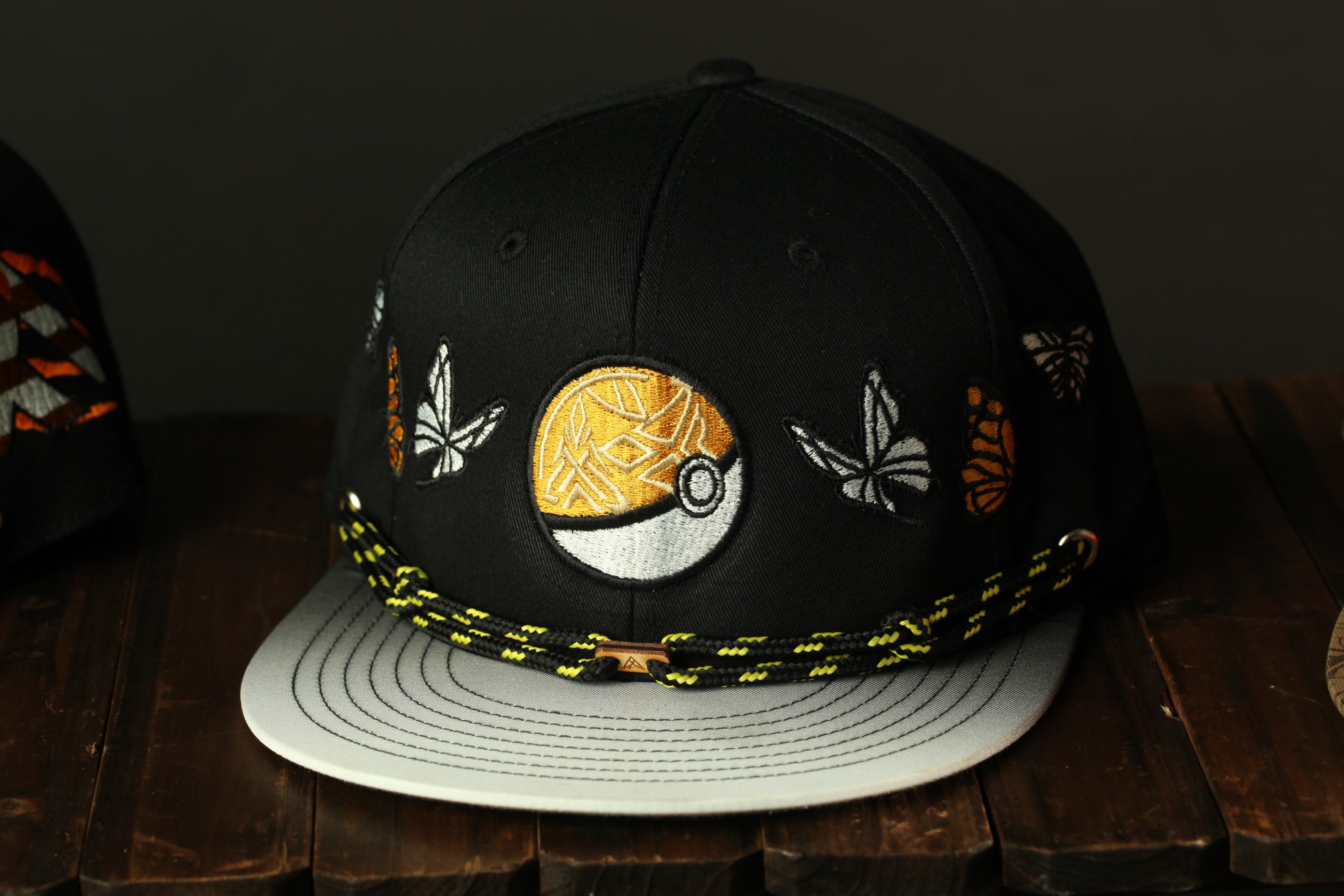 A black baseball cap with a gold and white Pokemon design, featuring a circular patch with a Pokemon ball and butterflies, is displayed on a wooden surface.