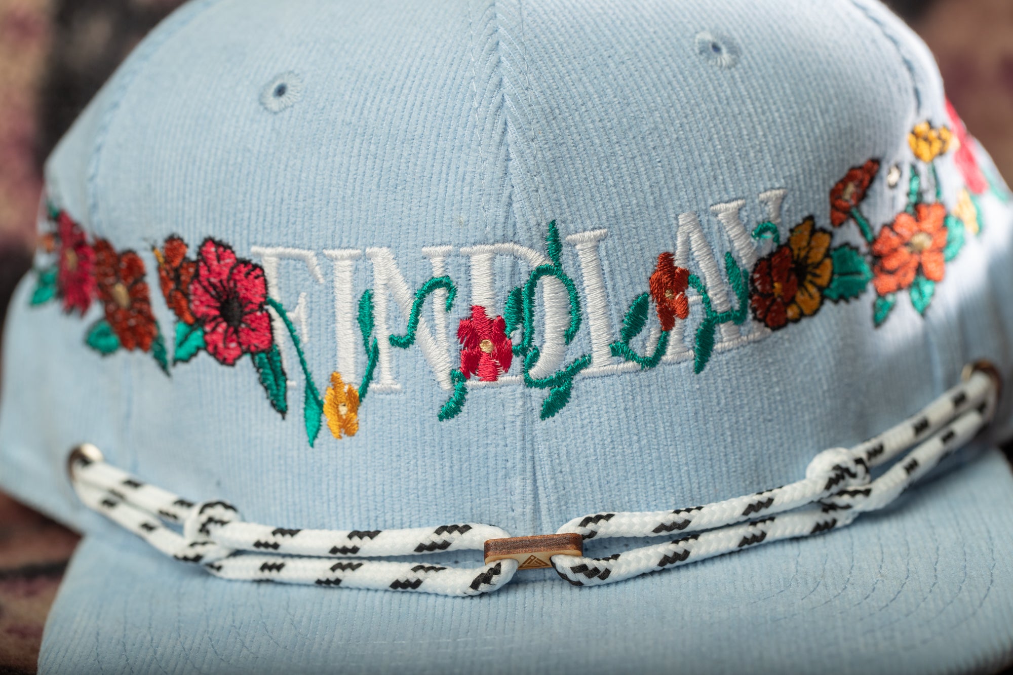 A close-up image of a light blue baseball cap with colorful floral embroidery and a black and white rope strap.