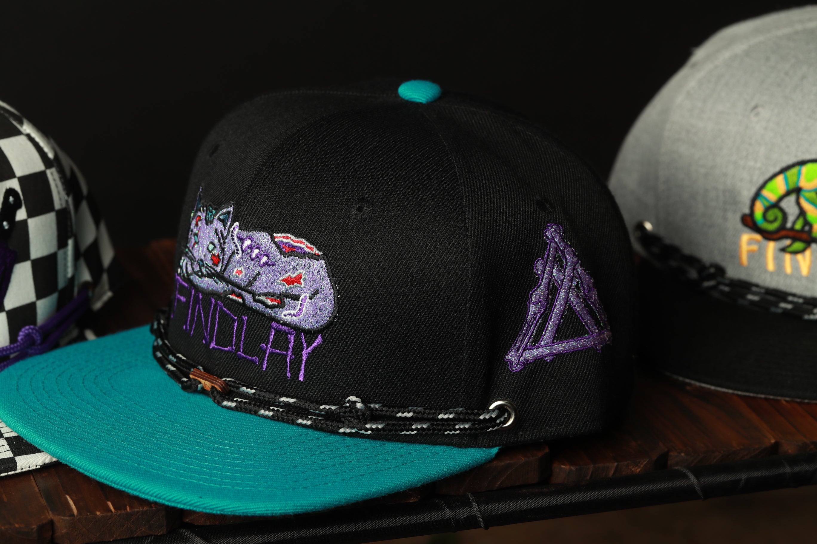 A close-up image of a black baseball cap with a teal brim and purple text, displayed on a wooden surface.