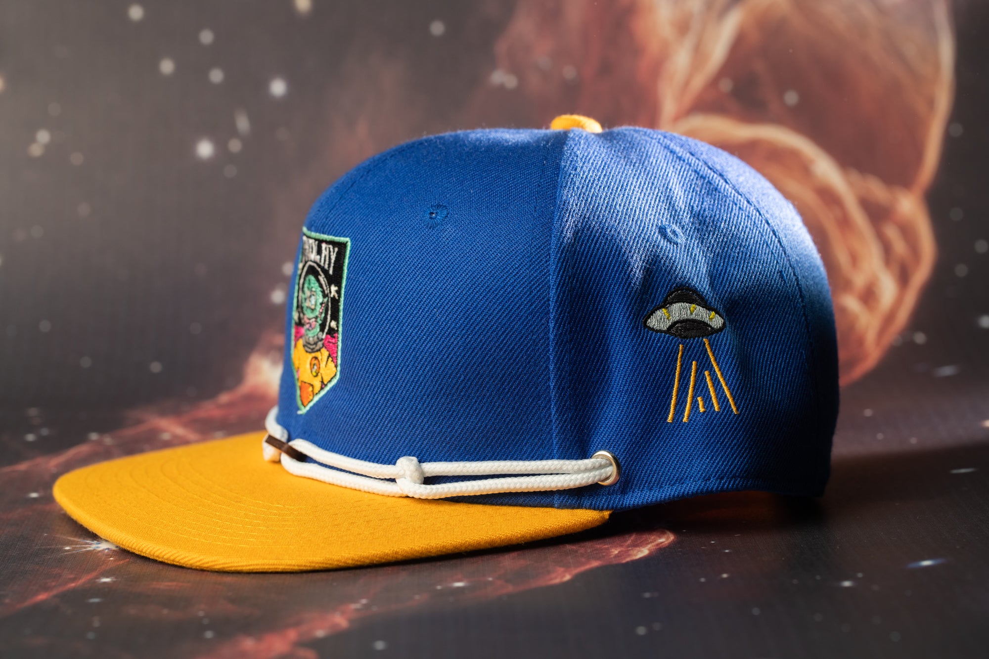 A blue baseball cap with a yellow brim and a small alien-like design on the side, set against a dark background with stars.
