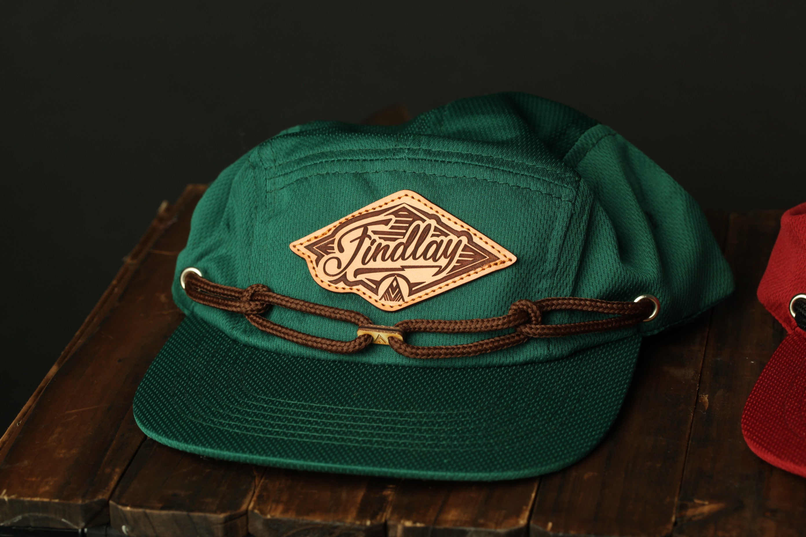 A green baseball cap with a brown leather patch that says "Findlay" on it, placed on a wooden surface.