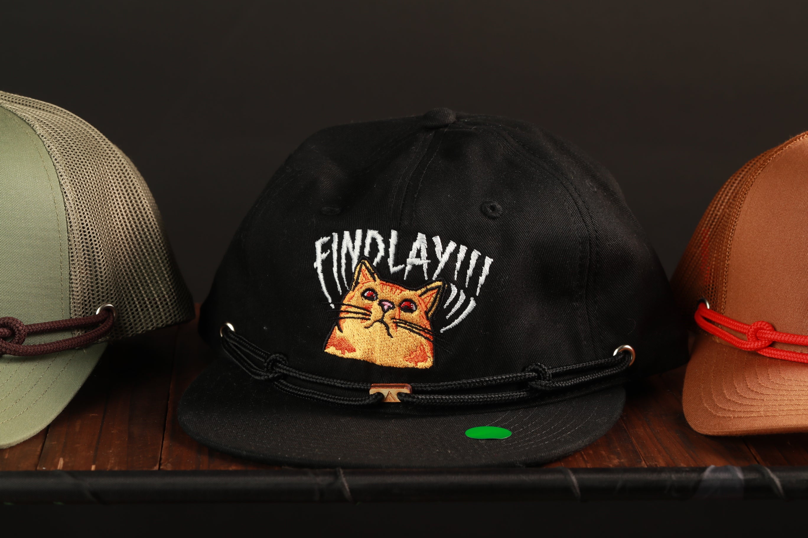 A black baseball cap with a cat embroidered on it, along with two other hats, is displayed on a wooden surface.