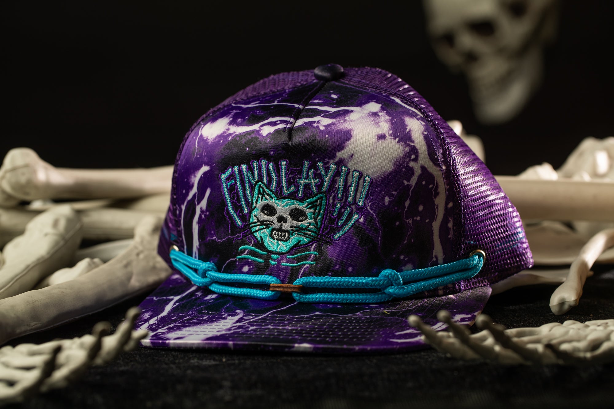 A purple baseball cap with a cat design and the text "FINOLAY" is displayed on a table, surrounded by skeletal bones.