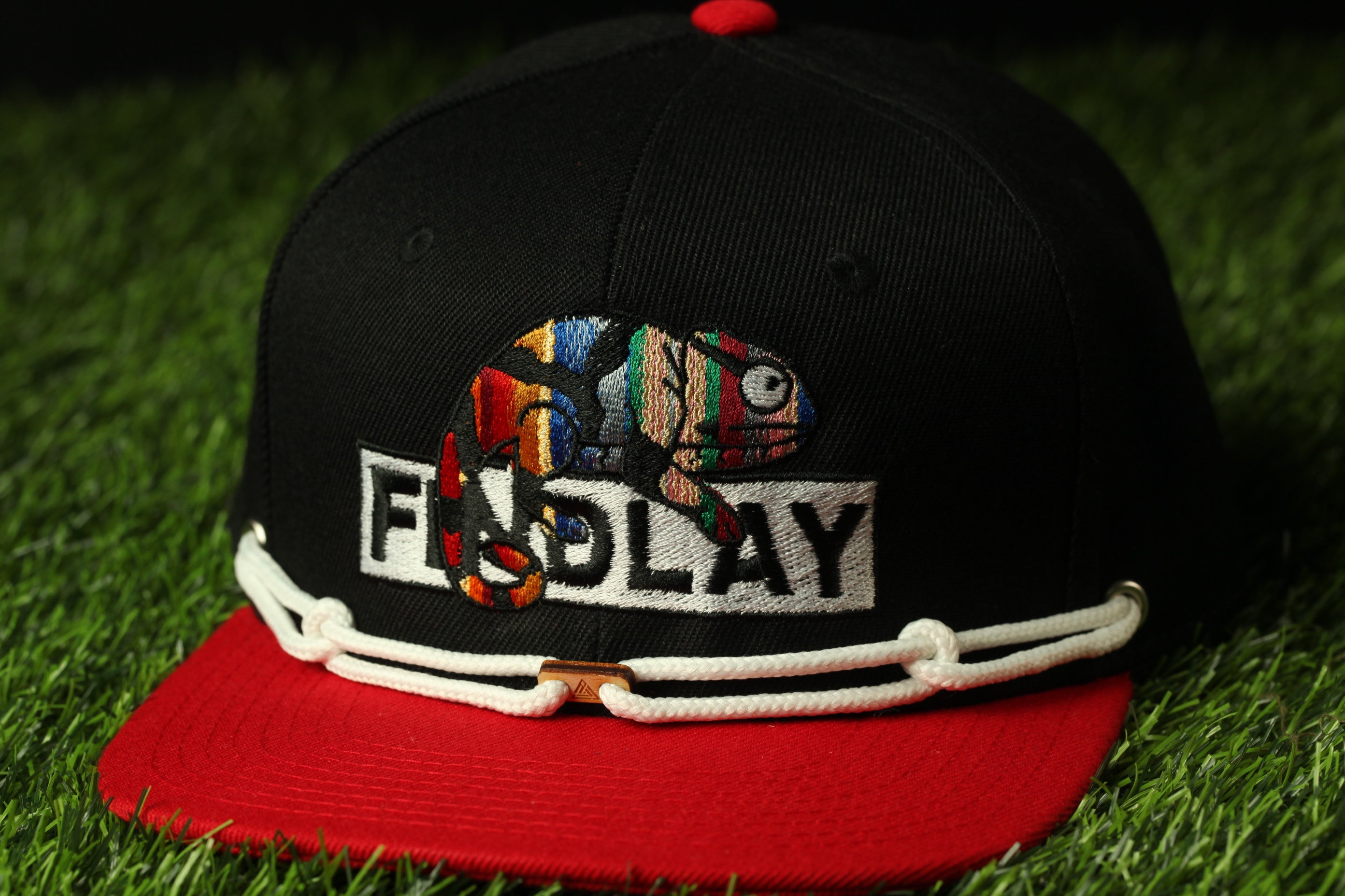 A black baseball cap with a red brim and a colorful chameleon design on the front, sitting on a grassy surface.