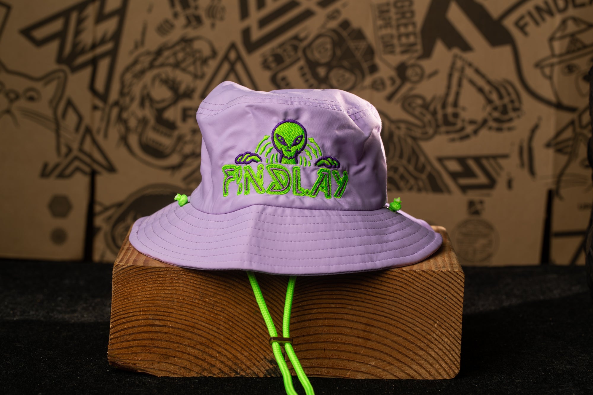 A purple bucket hat with a green alien design and the word "FINSLAY" printed on it, sitting on a wooden block.