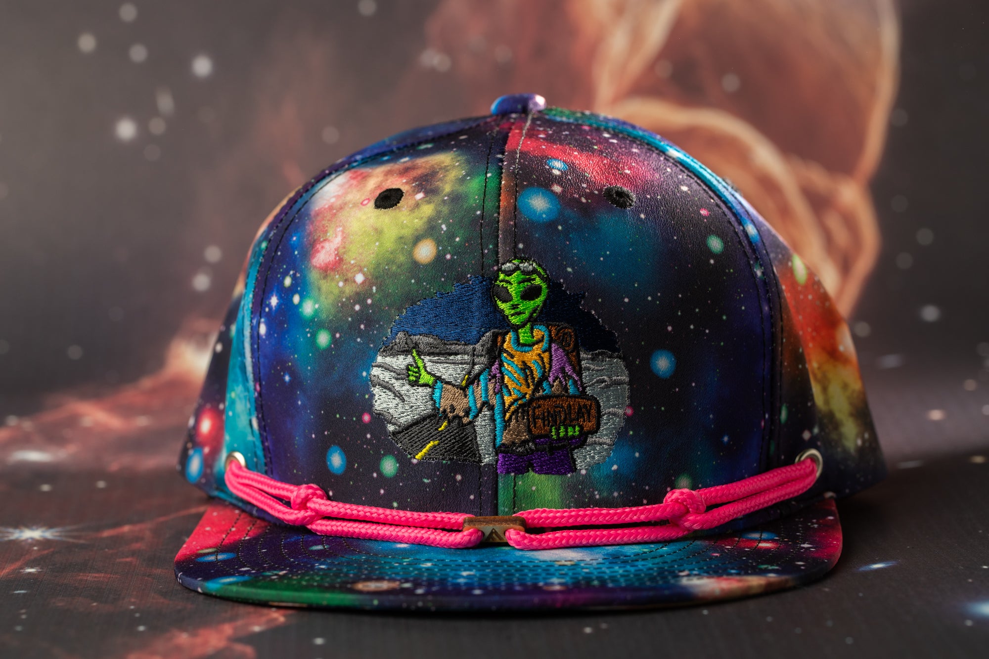 A colorful, patterned baseball cap with a space-themed design, featuring an alien character and a cosmic background.