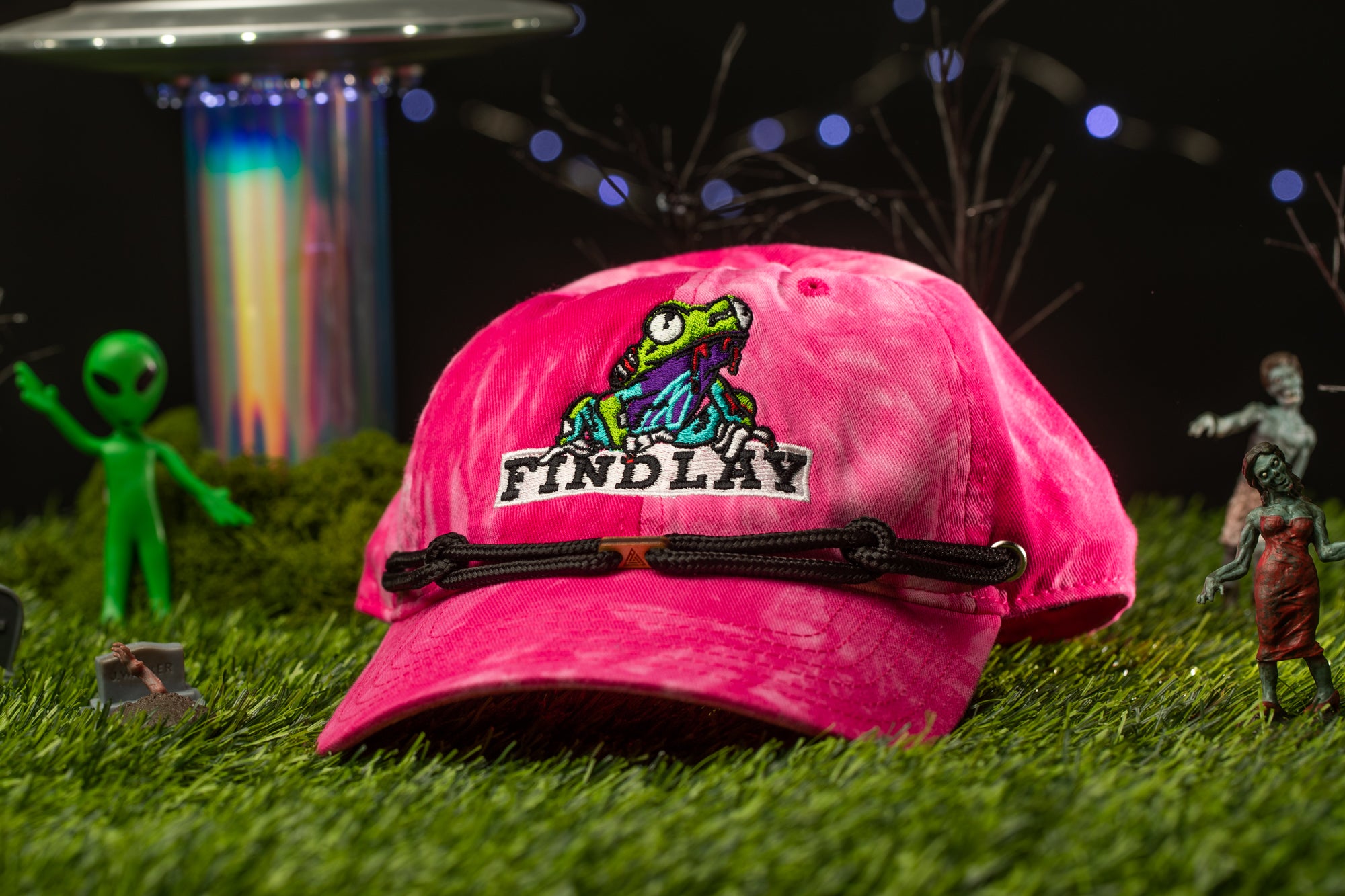 A pink baseball cap with a cartoon frog logo, placed on a grassy surface with various small toy figures and a colorful alien-like object in the background.