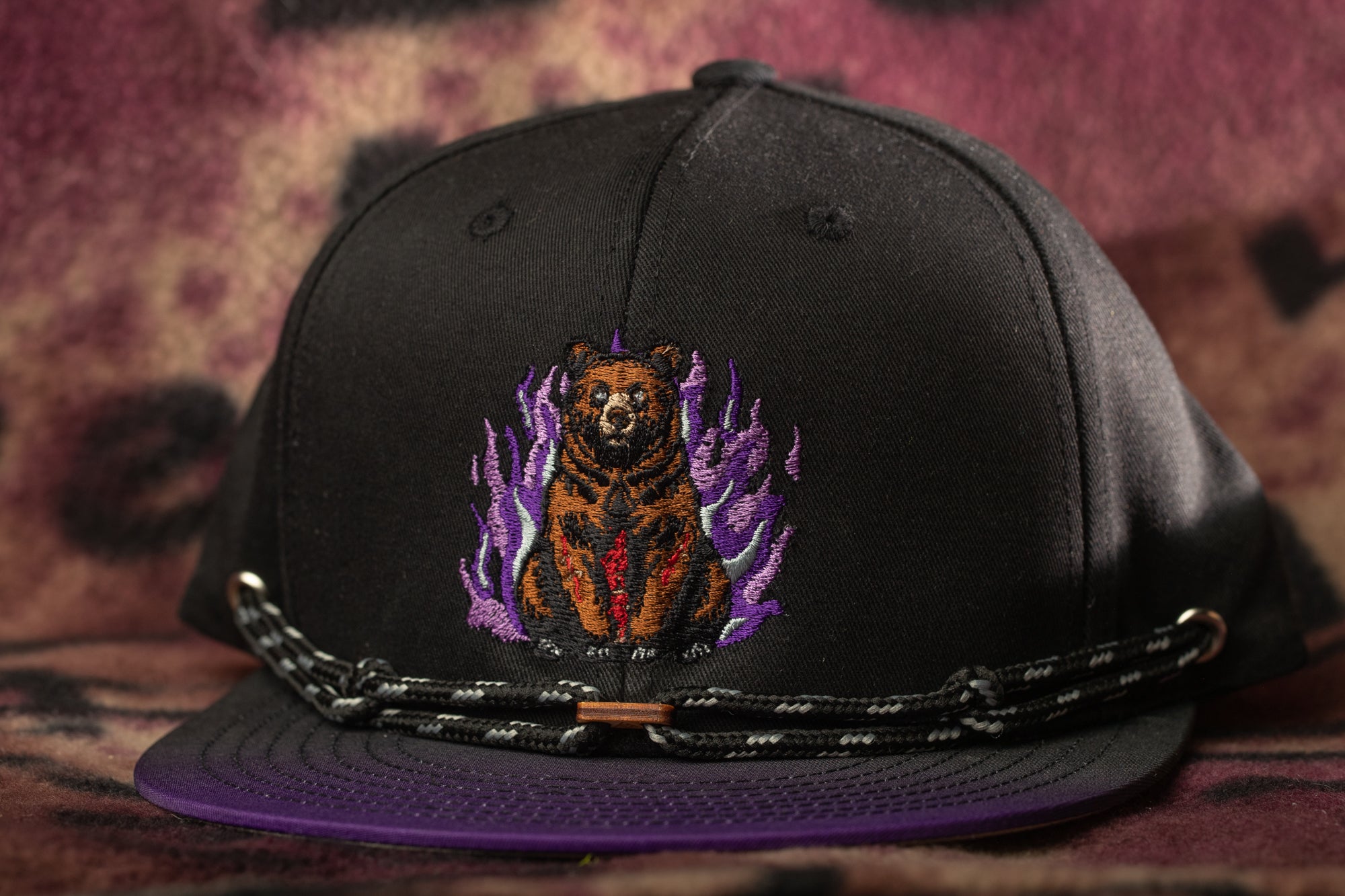 A black baseball cap with a purple and black design featuring a bear on a flame background.