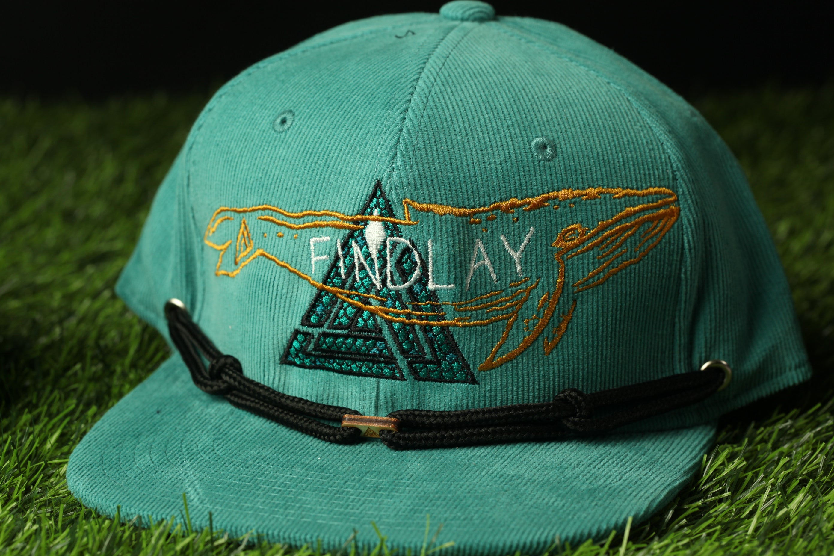 A turquoise baseball cap with a black strap and the word "FINDlay" embroidered on it, sitting on a grassy surface.