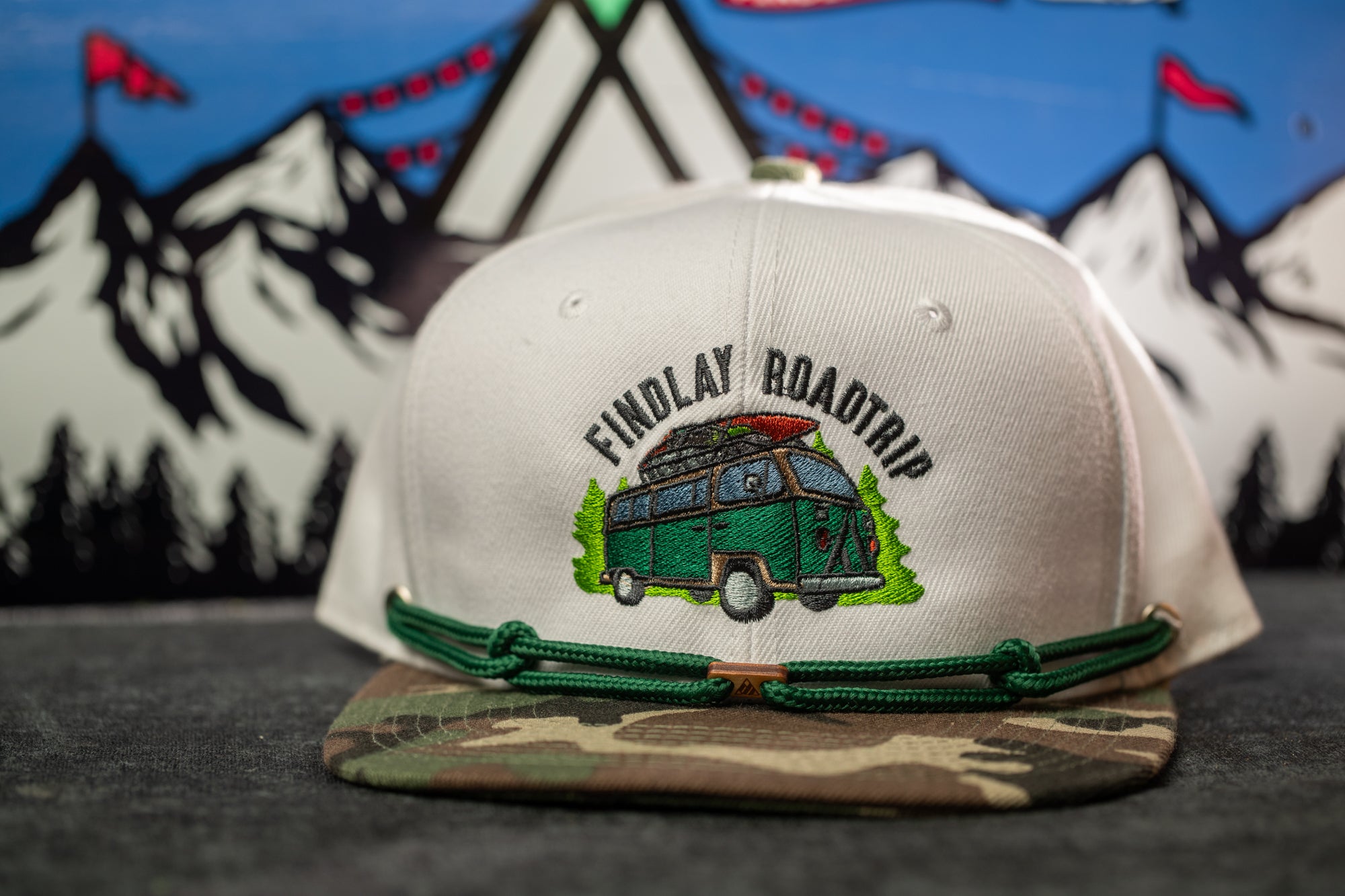 A white baseball cap with a camouflage pattern and a graphic of a van on it, with a mountain landscape in the background.